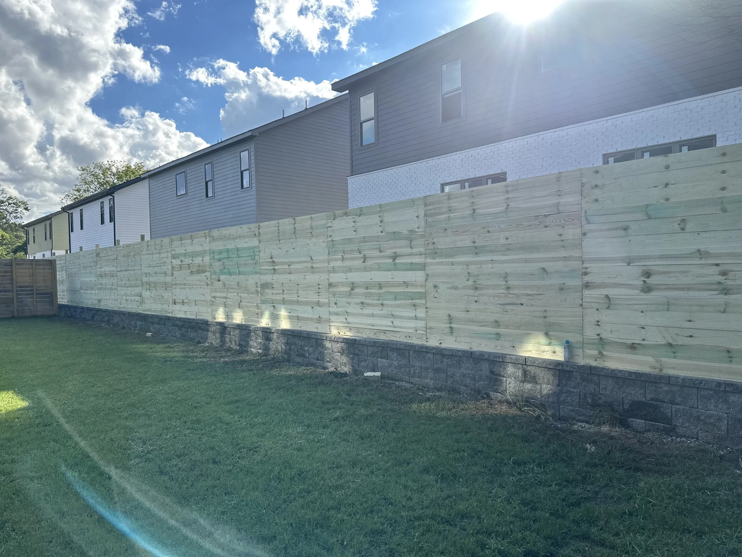 Backyard with a newly built wooden fence, green grass, and houses in the background under a partly cloudy sky with sunshine.