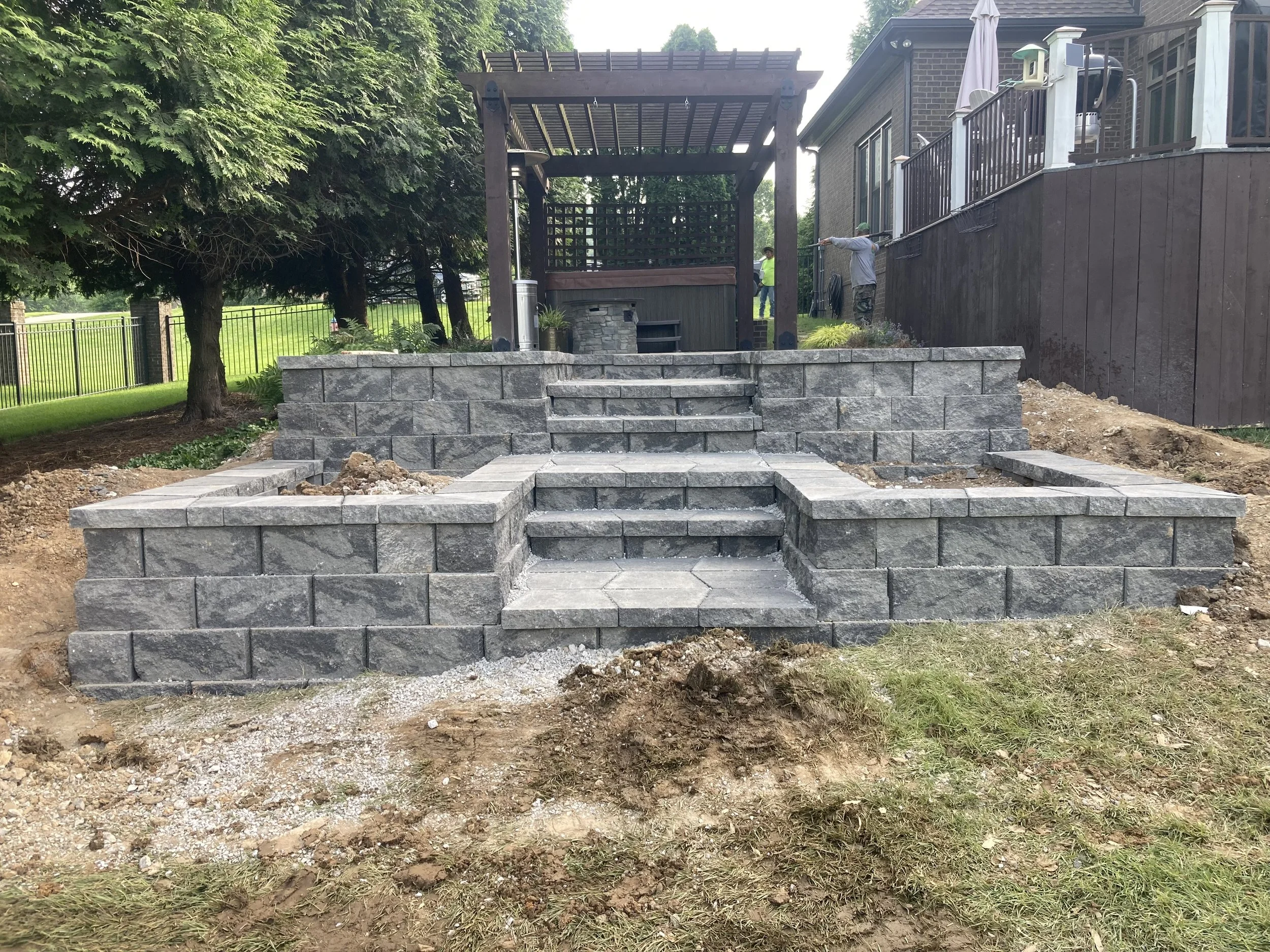 A backyard with a newly built stone staircase leading up to a wooden deck. The staircase has multiple levels with gray stone blocks, and the surrounding area is still under construction with dirt and gravel. There are two people working in the backgr