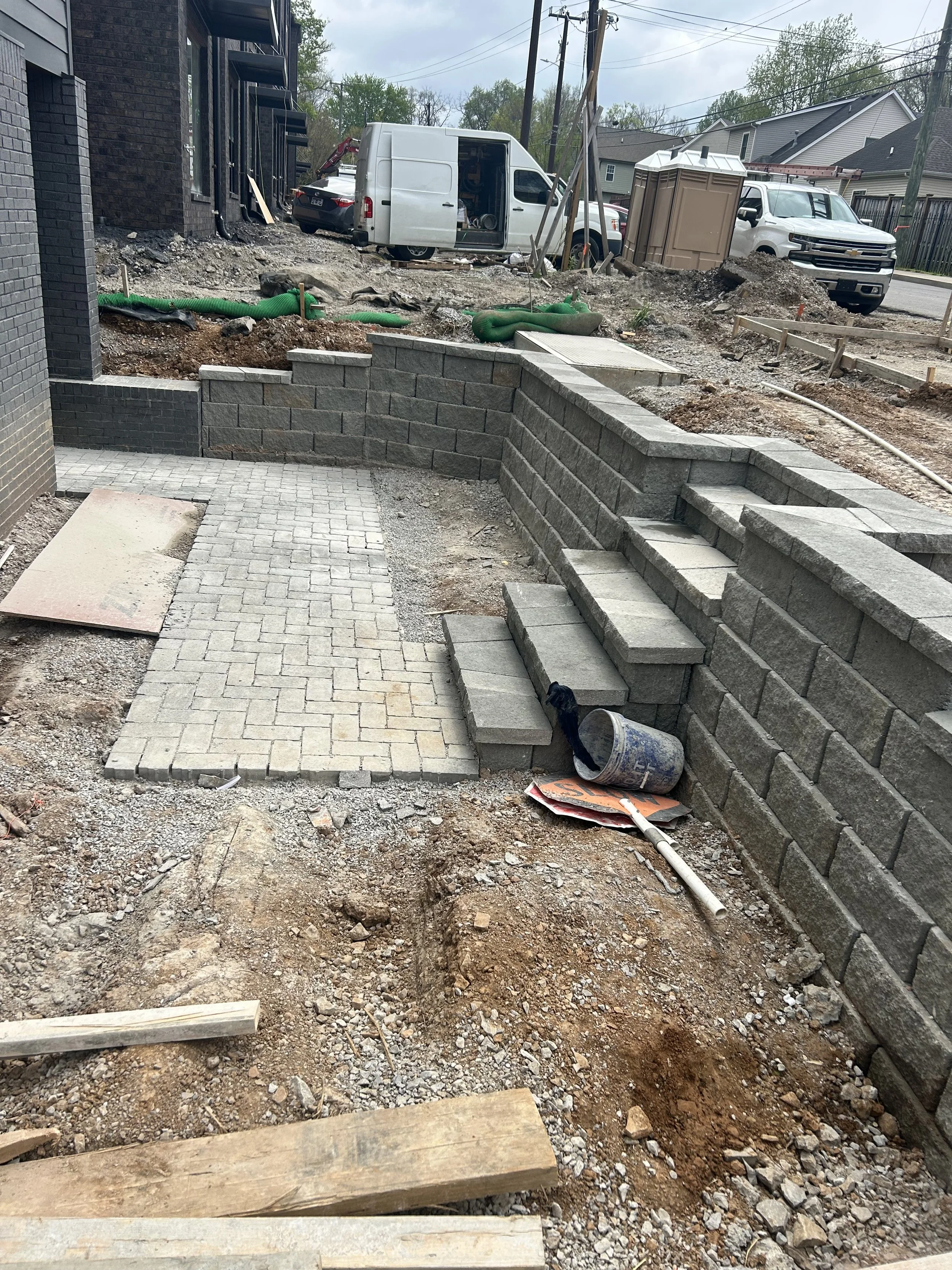Construction site with brick paver walkway, retaining wall, and ongoing landscaping, with trucks and construction tools visible in the background.