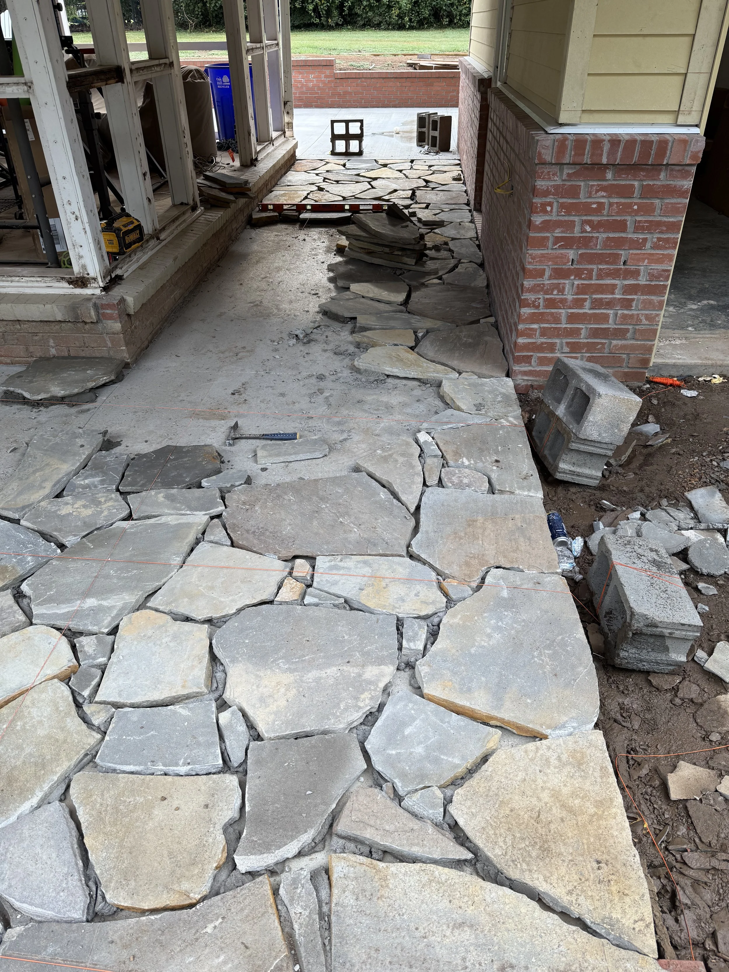 Outdoor project in progress with a partially laid stone pathway, construction tools, and loose stones and cinder blocks, near a house with brick and siding exterior.