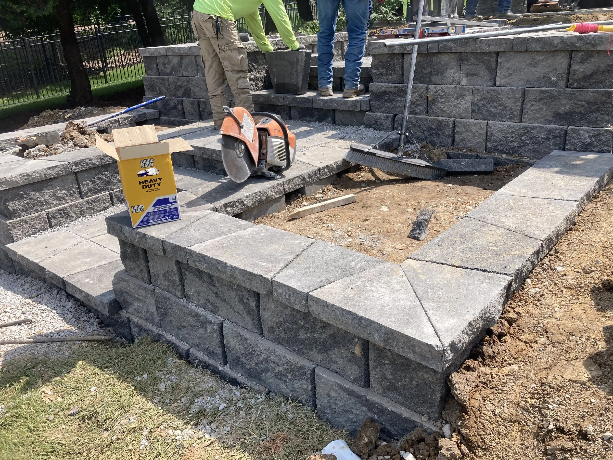 Workers building a stone patio with layered stone blocks, construction tools, and materials visible, with a fence and trees in the background.