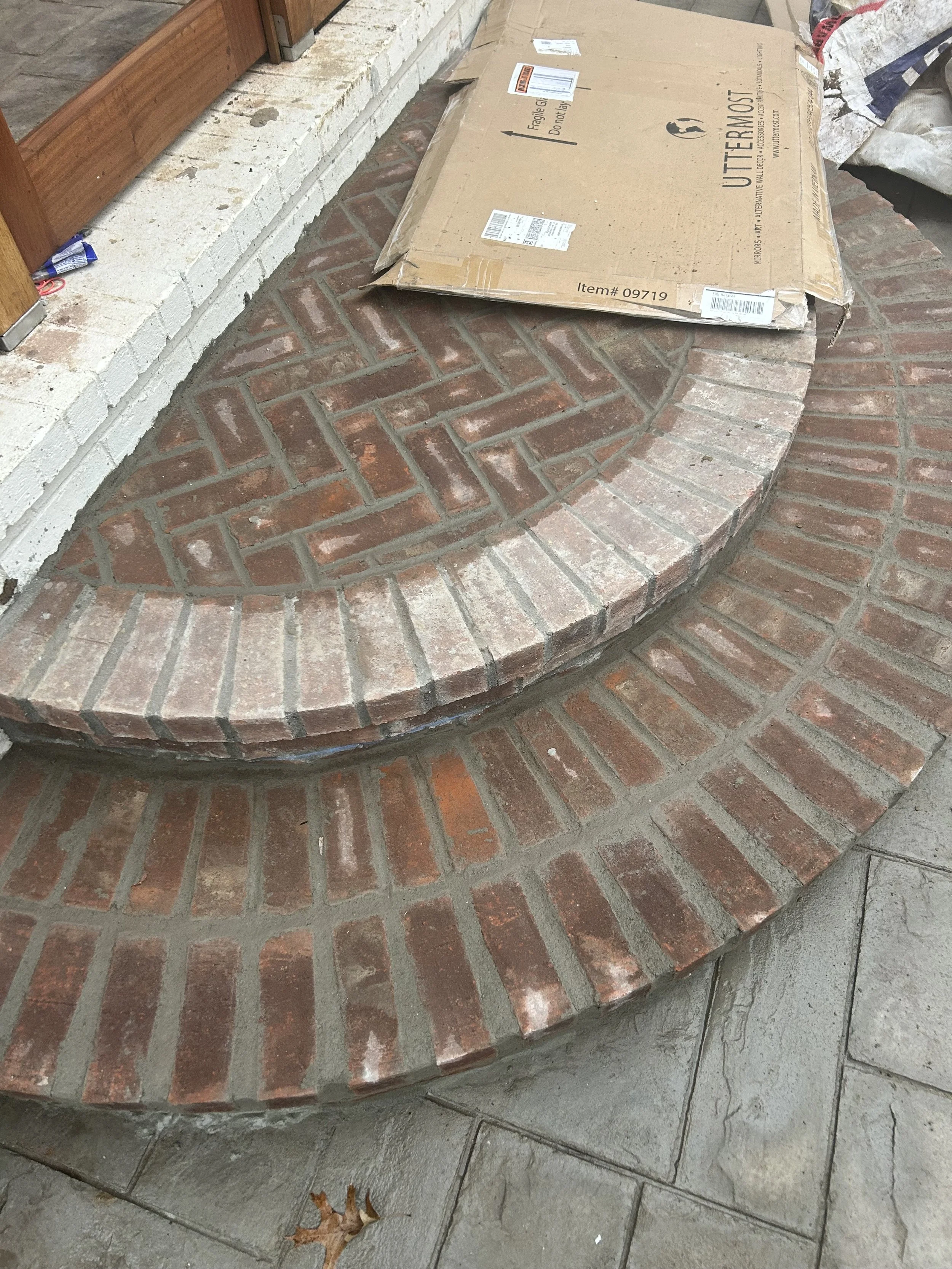 Close-up view of a brick porch with a semi-circular step, a cardboard box on top, and a small fallen leaf on the concrete. The brickwork shows both new and weathered bricks.