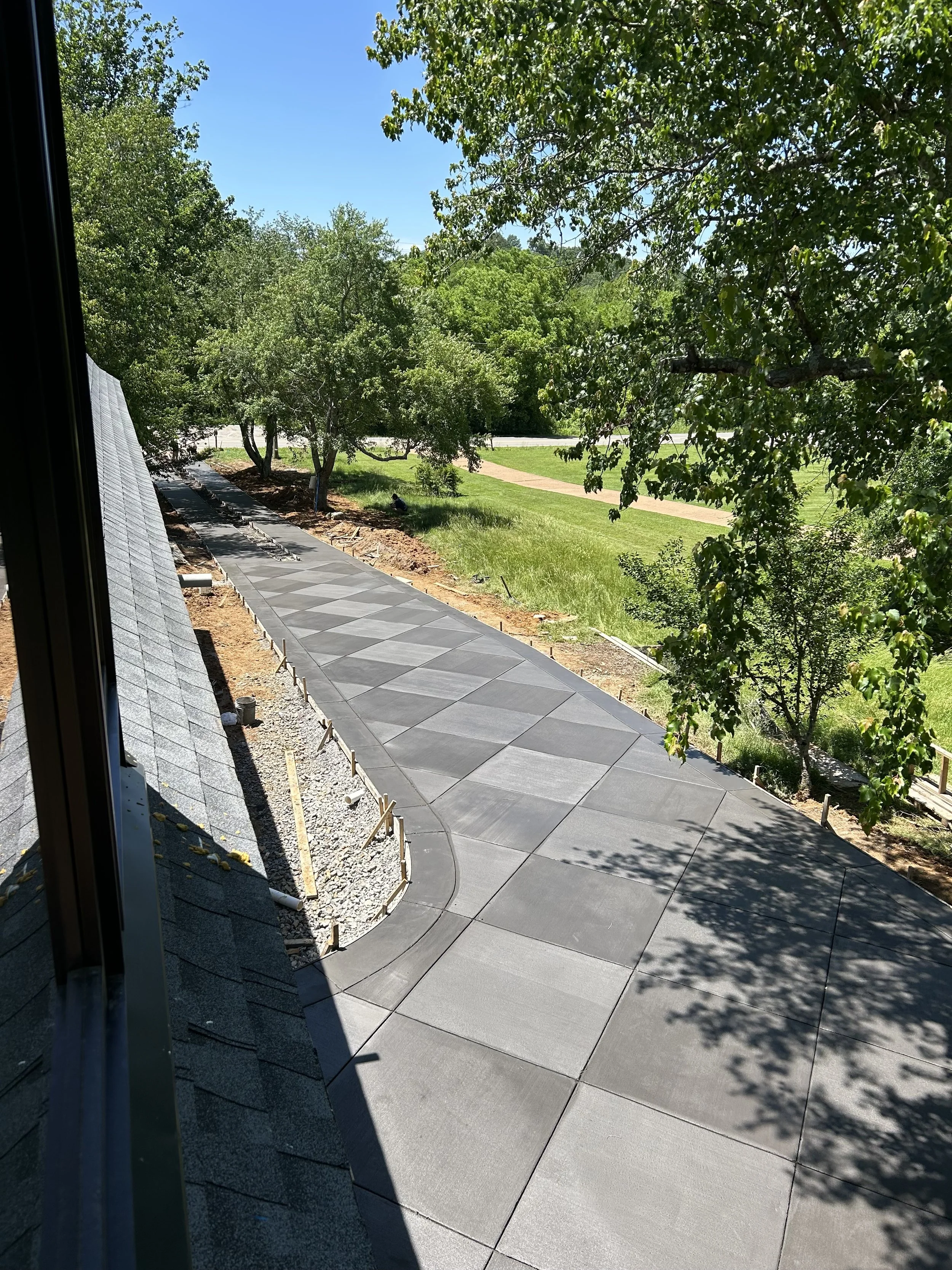 View of a newly paved sidewalk with large concrete slabs, surrounded by green trees and grass under a clear blue sky, with construction materials and outlines along the edge.