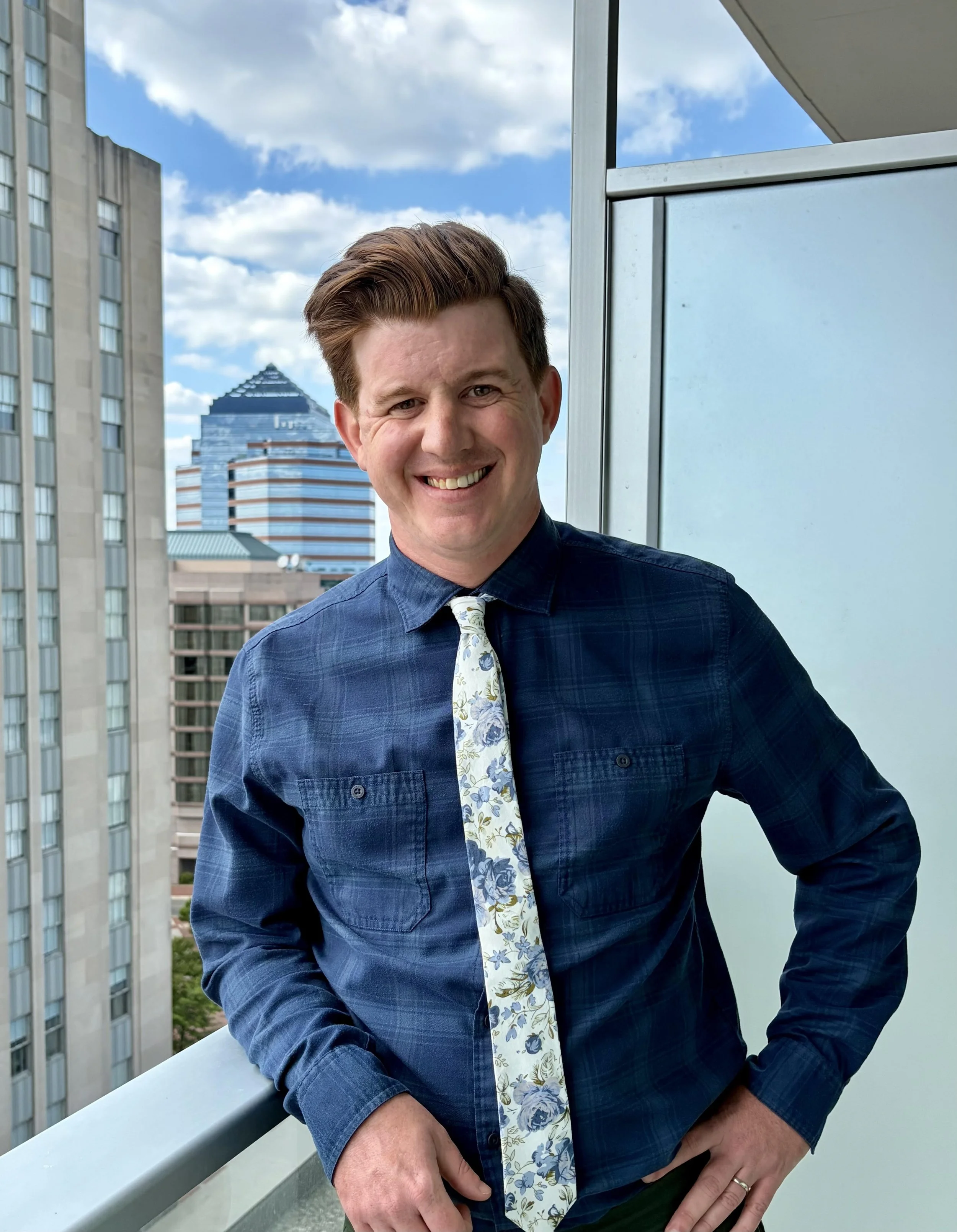 A man standing on a balcony with city buildings in the background. He's smiling, wears a blue plaid shirt, a floral tie, and has short, styled hair.