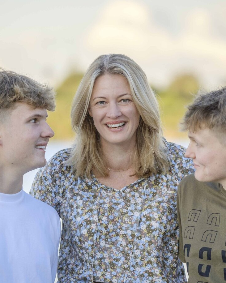 A woman in a floral blouse smiling with two boys on either side, outdoors with blurred trees in the background.