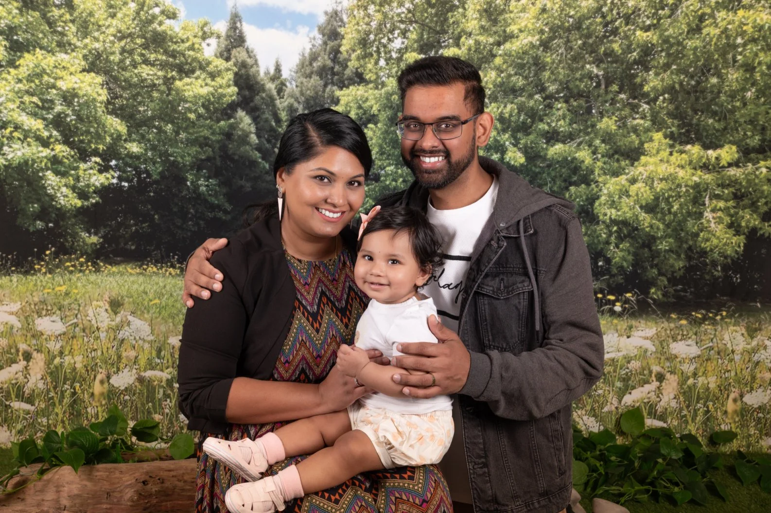 Family of three smiling and posing outdoors with green trees and yellow flowers in the background. 
