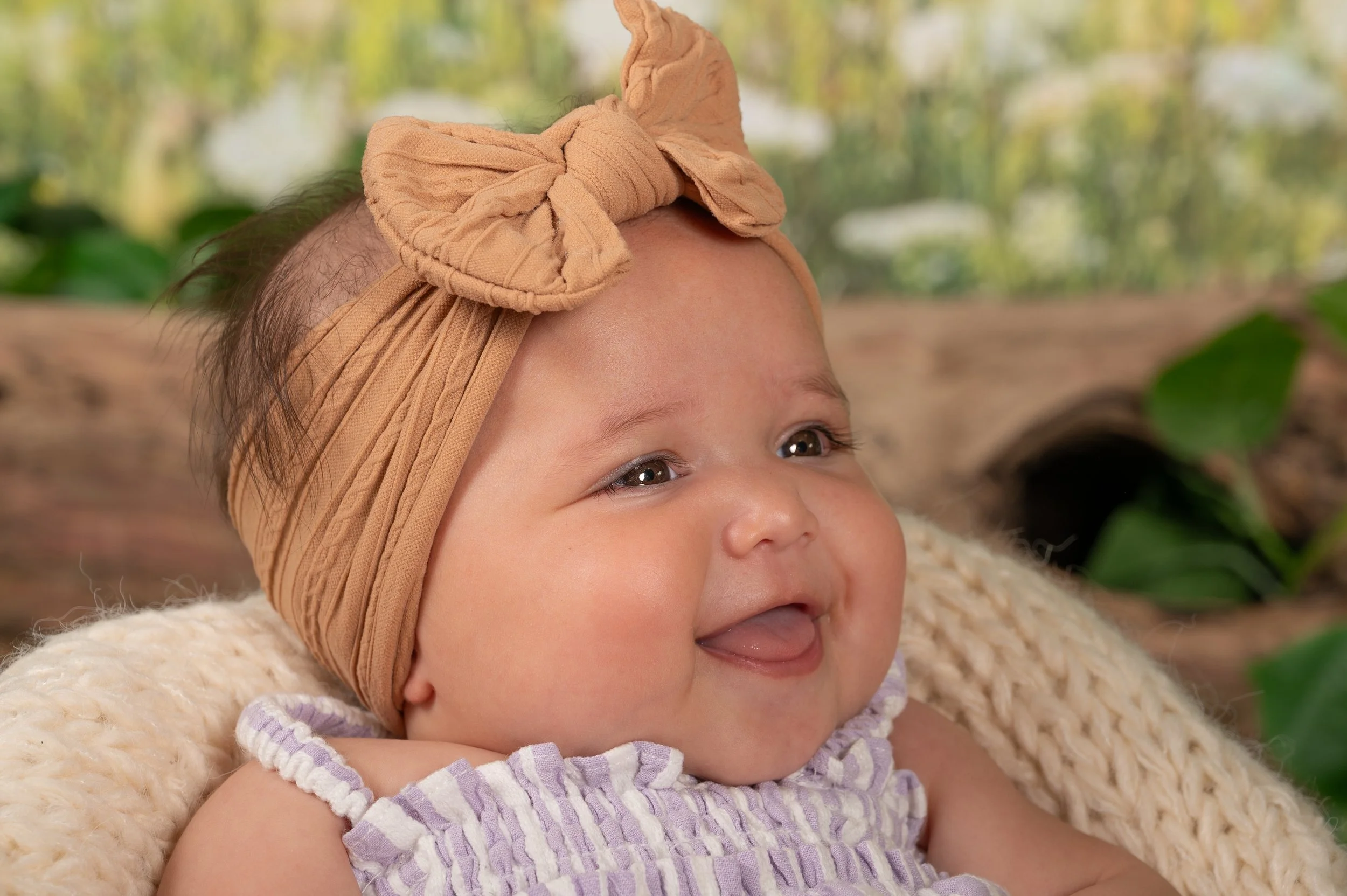 Close-up of a smiling baby girl with a beige headband and a purple striped dress, lying on a cream-colored blanket with foliage in the background.