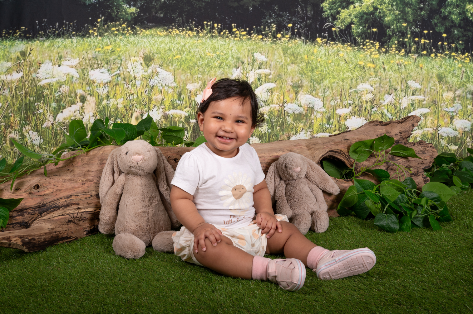 A smiling toddler girl sitting on grass in front of a backdrop of a field with white flowers.