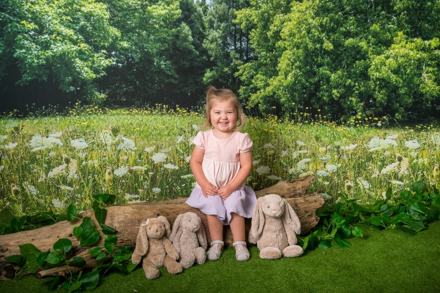 A young girl with blonde hair sitting on a log in a forest-themed backdrop.