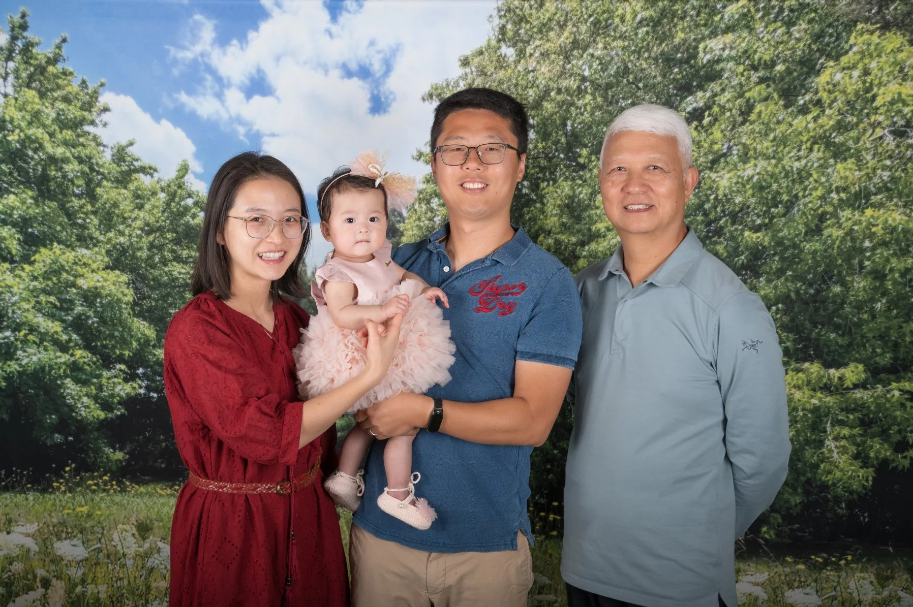 A family of four standing outdoors in front of a scenic background with trees and blue sky.