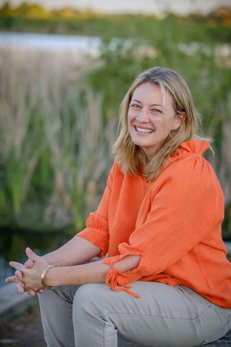 A woman sitting outdoors near water and green plants, smiling at the camera, wearing an orange blouse.
