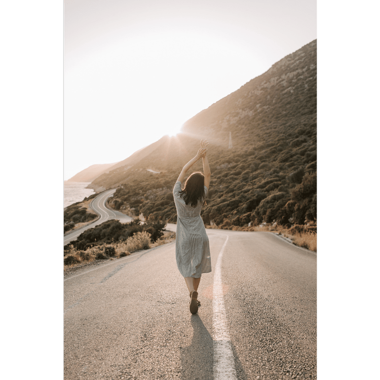 A woman walking down a sunlit coastal road with her arms raised