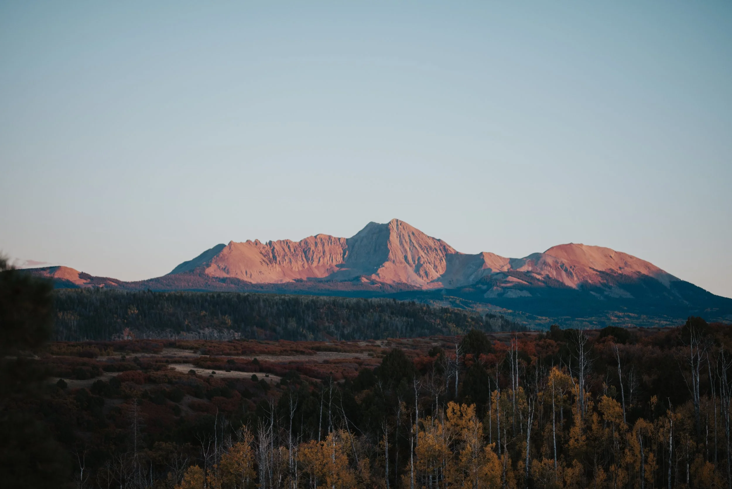 Mountain range at sunrise or sunset with a forest in the foreground and clear sky above.
