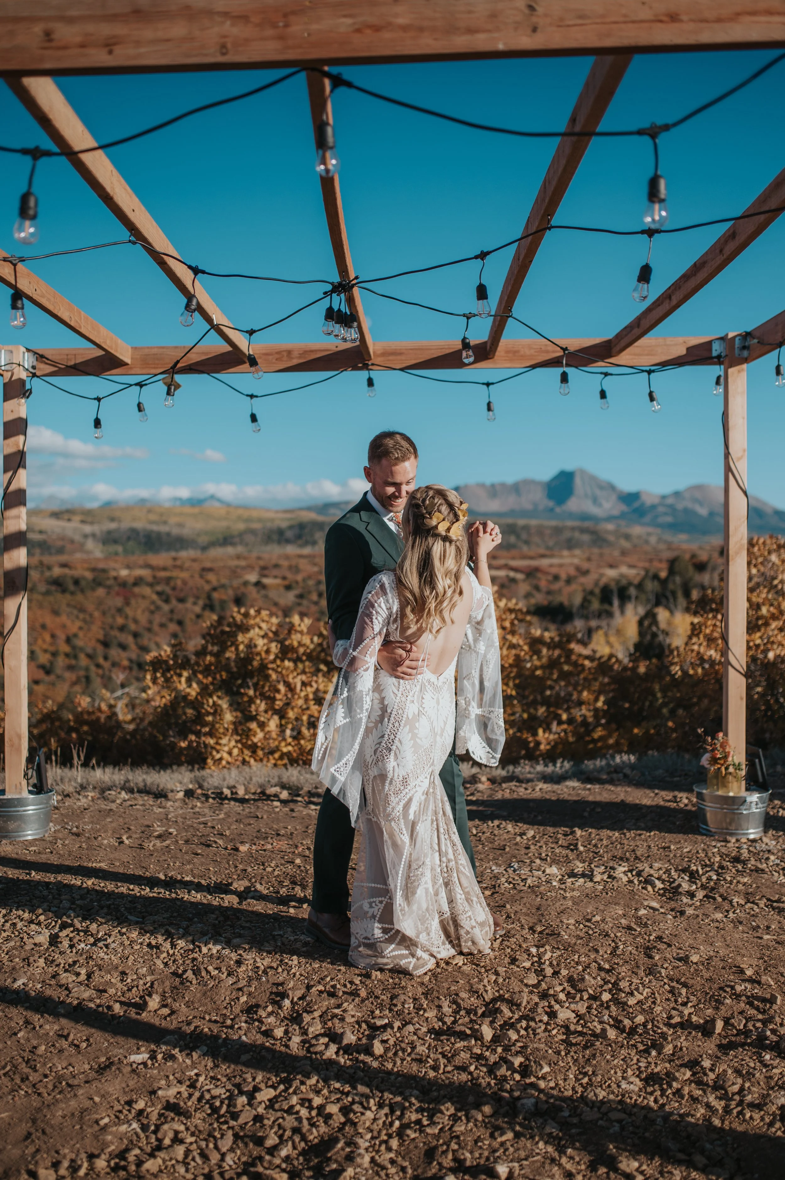 A bride and groom dancing outdoors under a wooden framework with string lights against a scenic mountain and autumn landscape.