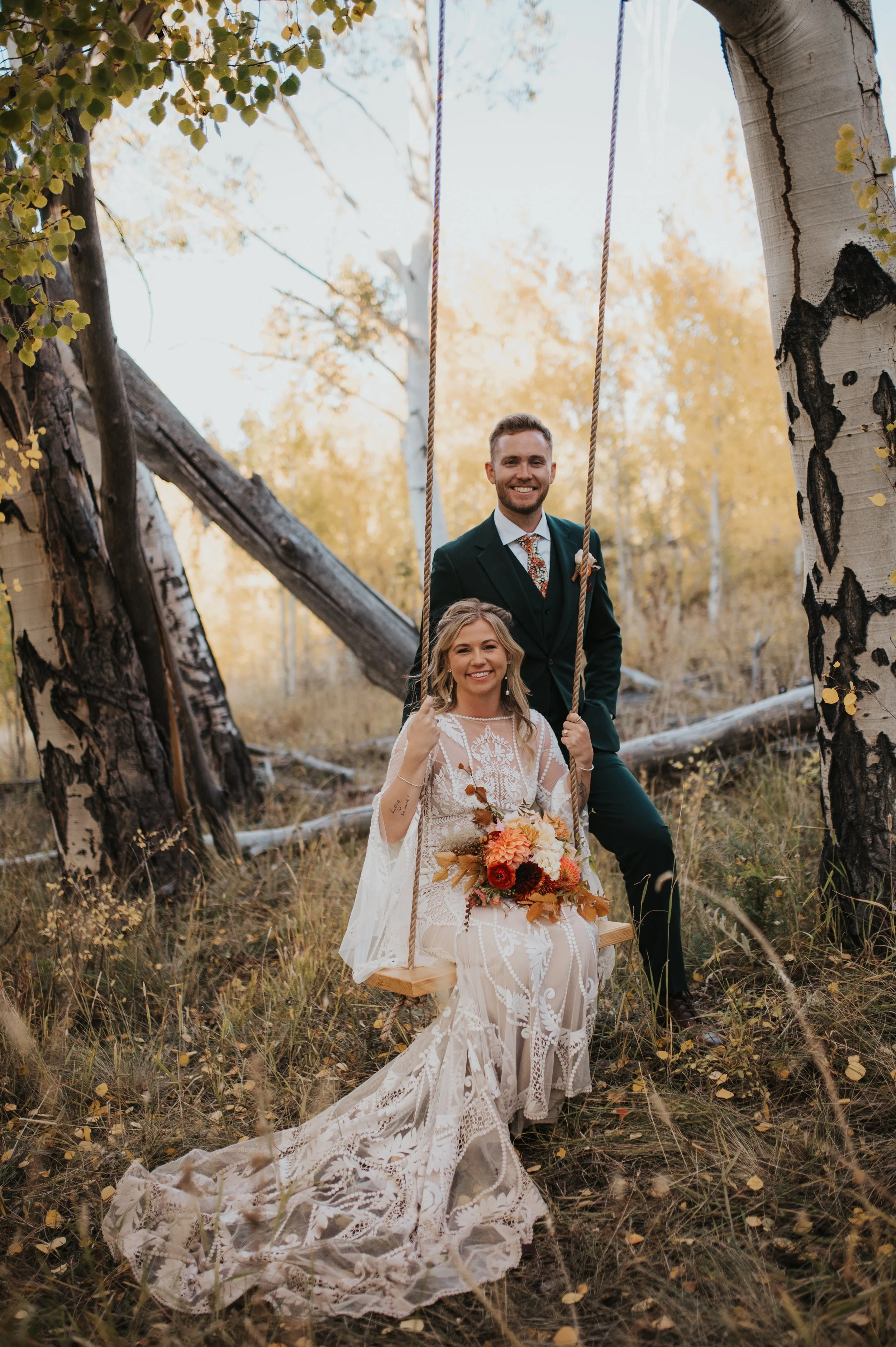 A couple is outdoors in a forest during autumn, with the woman sitting on a wooden swing and the man standing behind her. The woman is wearing a white lace wedding dress and holding a bouquet of flowers, while the man is dressed in a dark suit with a floral tie. They are smiling happily amidst fallen leaves and surrounded by trees with yellowing leaves.