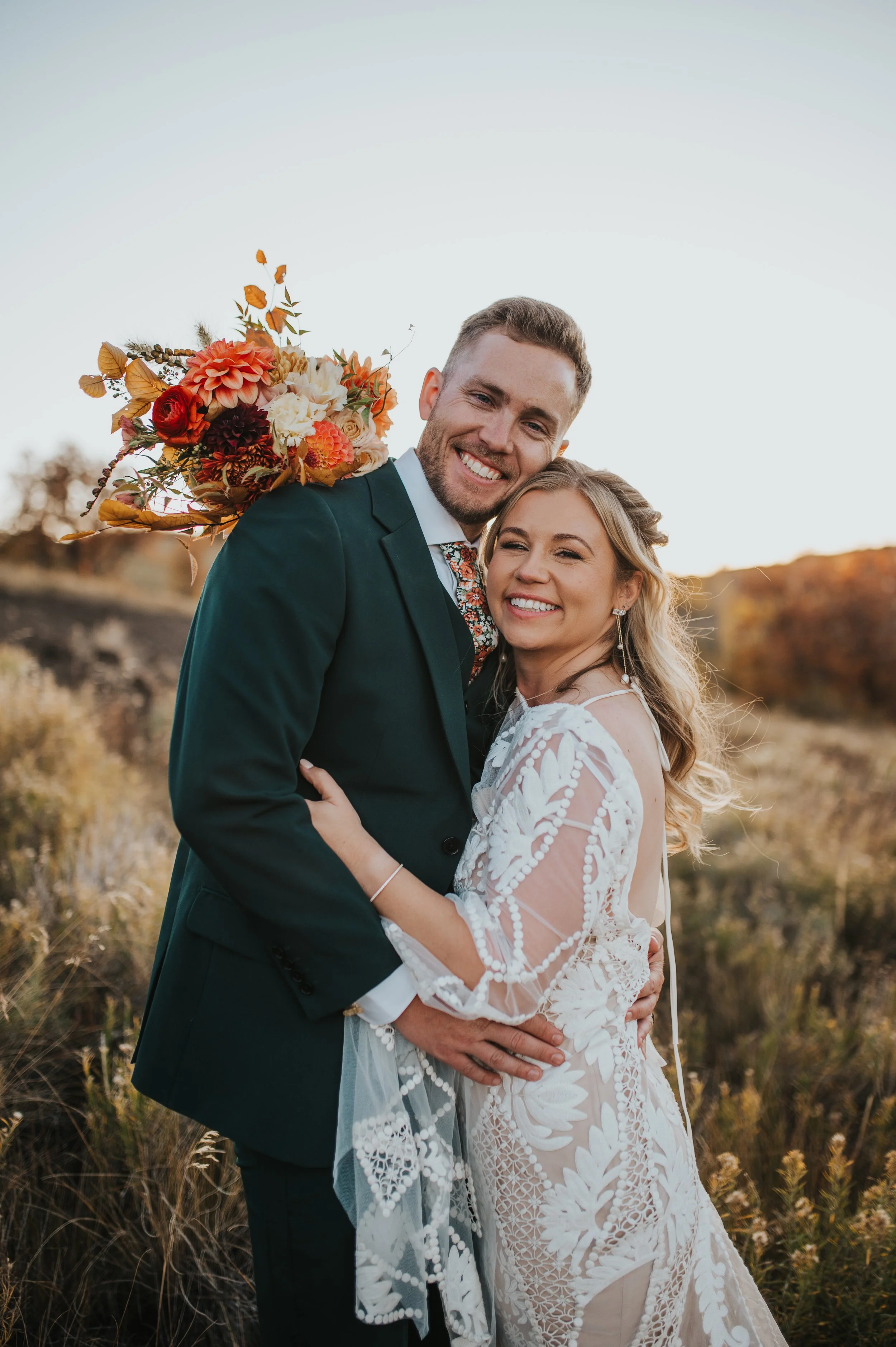 A smiling bride and groom hugging outdoors in a field during sunset, with the groom holding a bouquet of flowers over his shoulder.