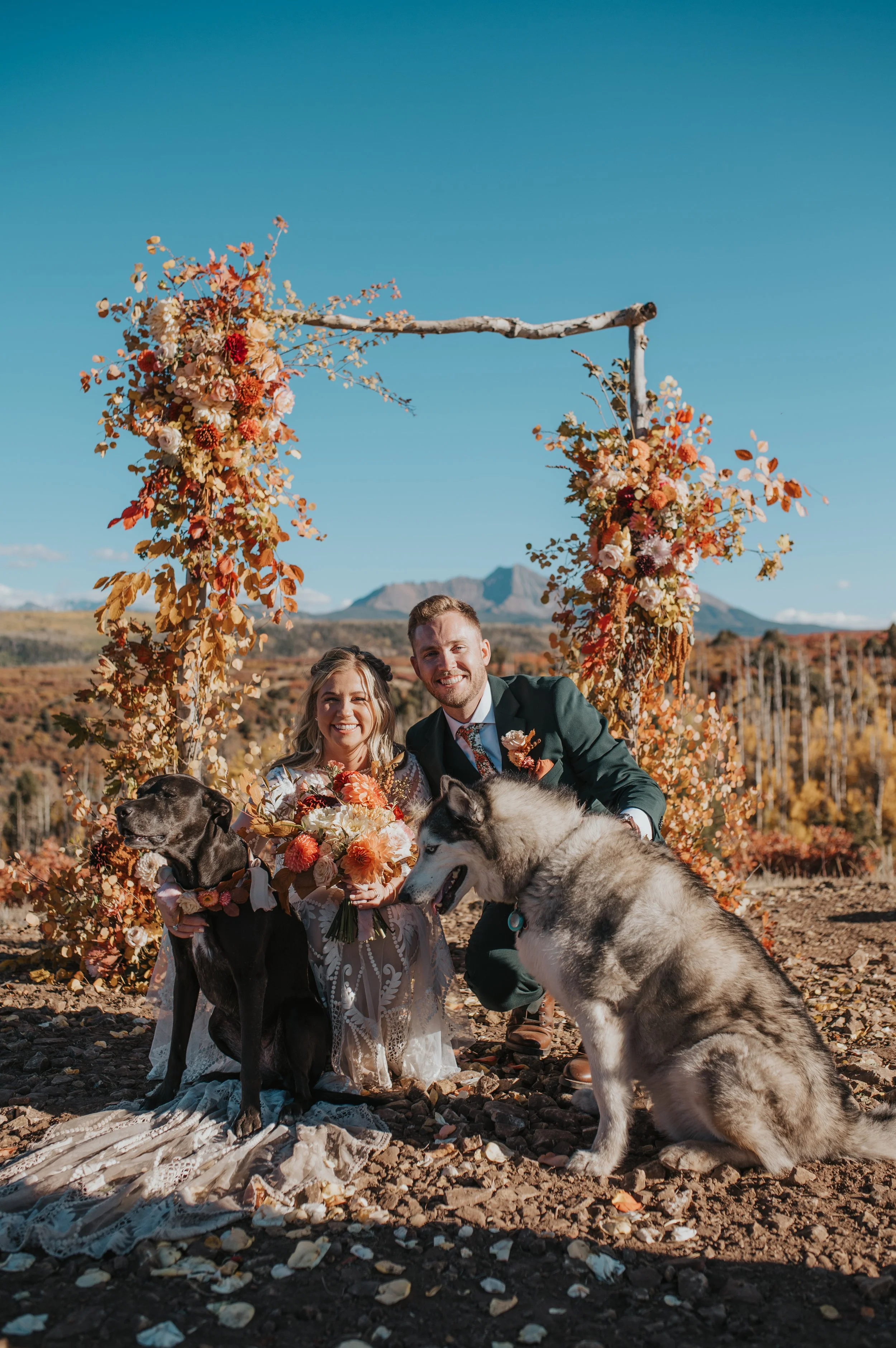A newlywed couple poses outdoors with their two dogs under an autumn-themed floral arch, with mountains in the background.