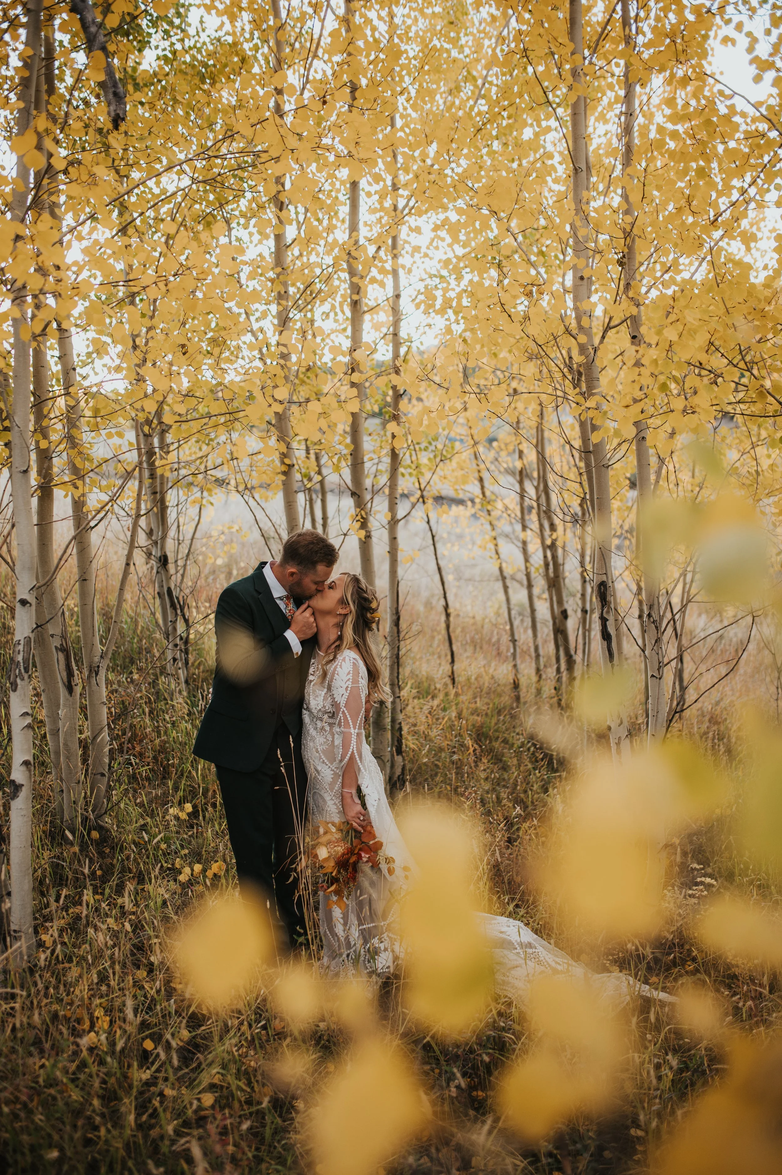 A bride and groom kissing in an autumn forest with yellow leaves and tall grasses.