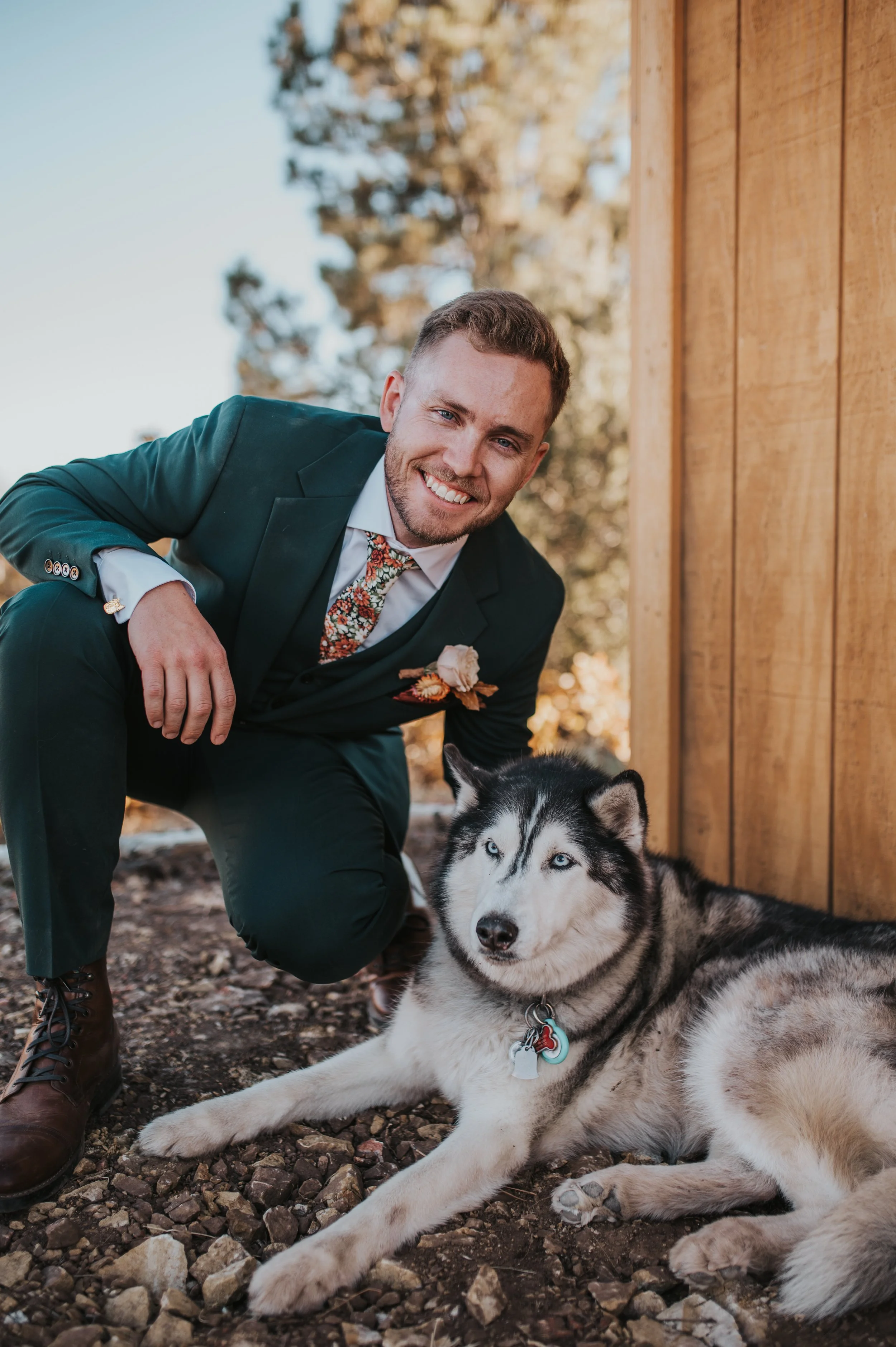 A man in a suit crouching beside a Siberian Husky dog outdoors, with a wooden wall in the background and trees.