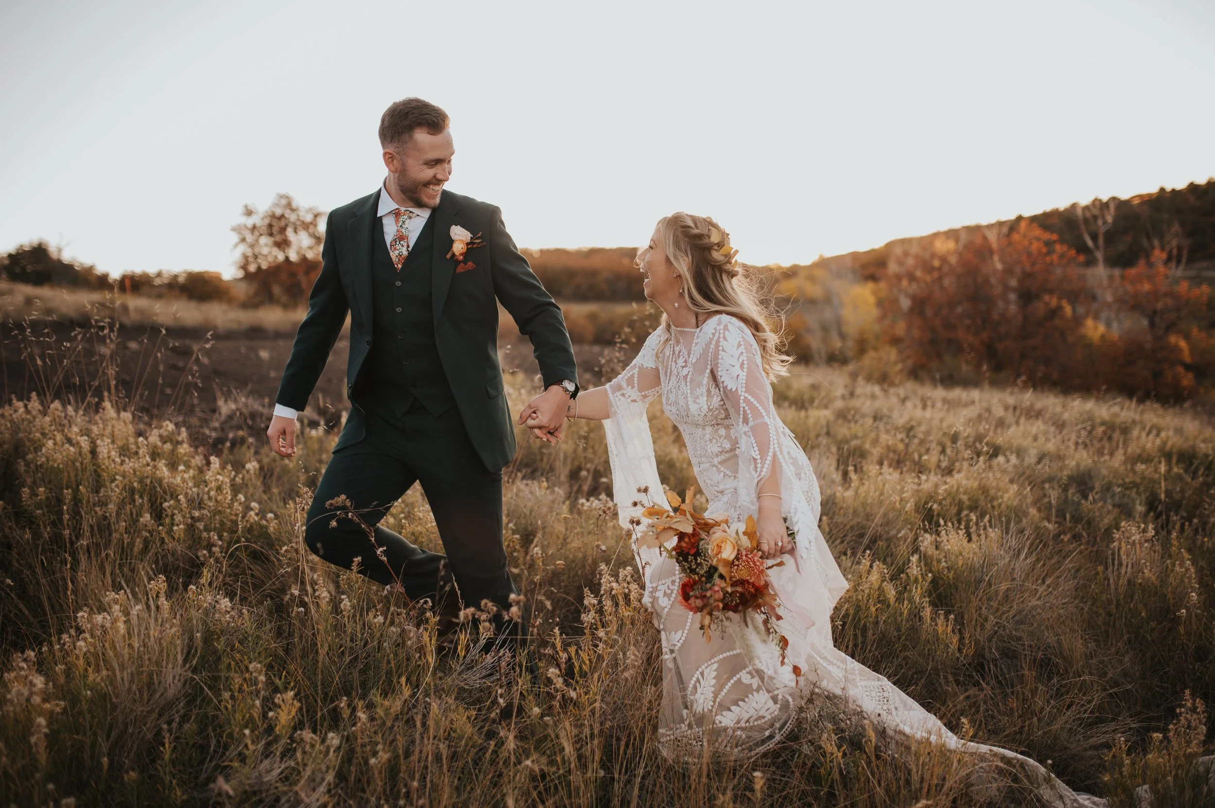 A happy couple in wedding attire holding hands in a field during autumn, with colorful trees and a setting sun in the background.