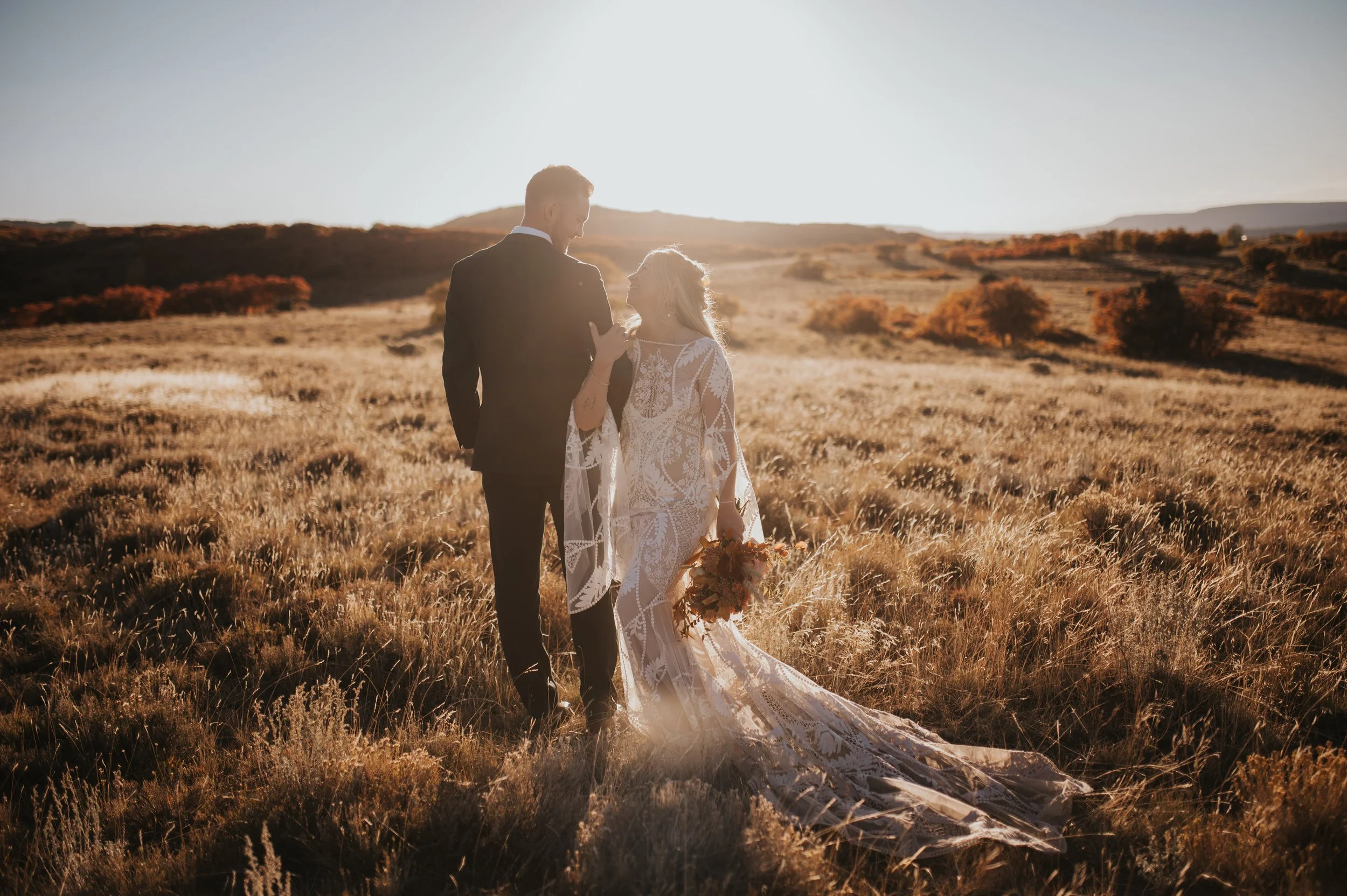 A bride and groom walking hand-in-hand through a field at sunset, with the bride holding a bouquet, during their wedding photos.