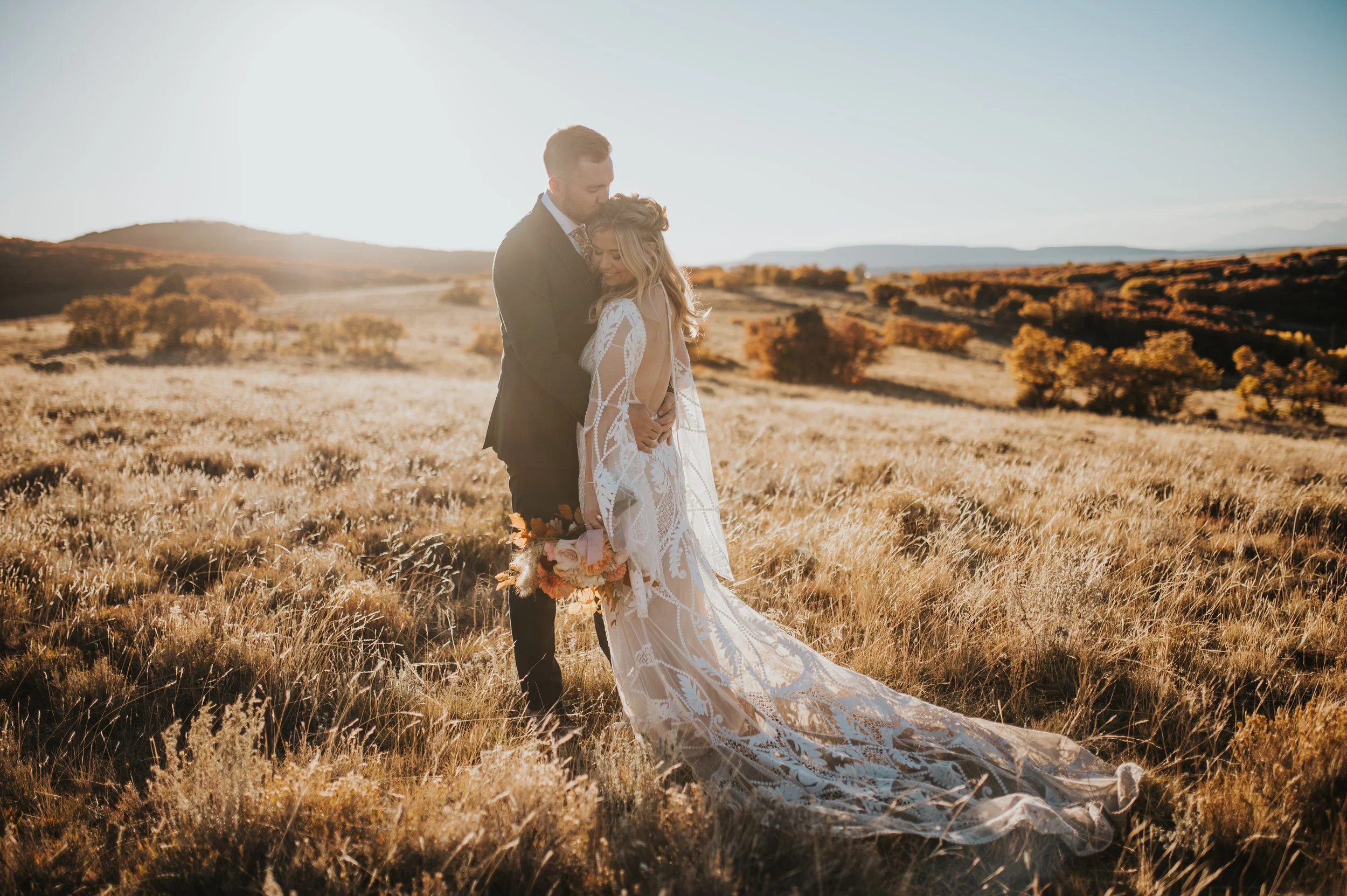 A couple in wedding attire standing in a grassy field during sunset, embracing each other with a scenic landscape of rolling hills and trees in the background.