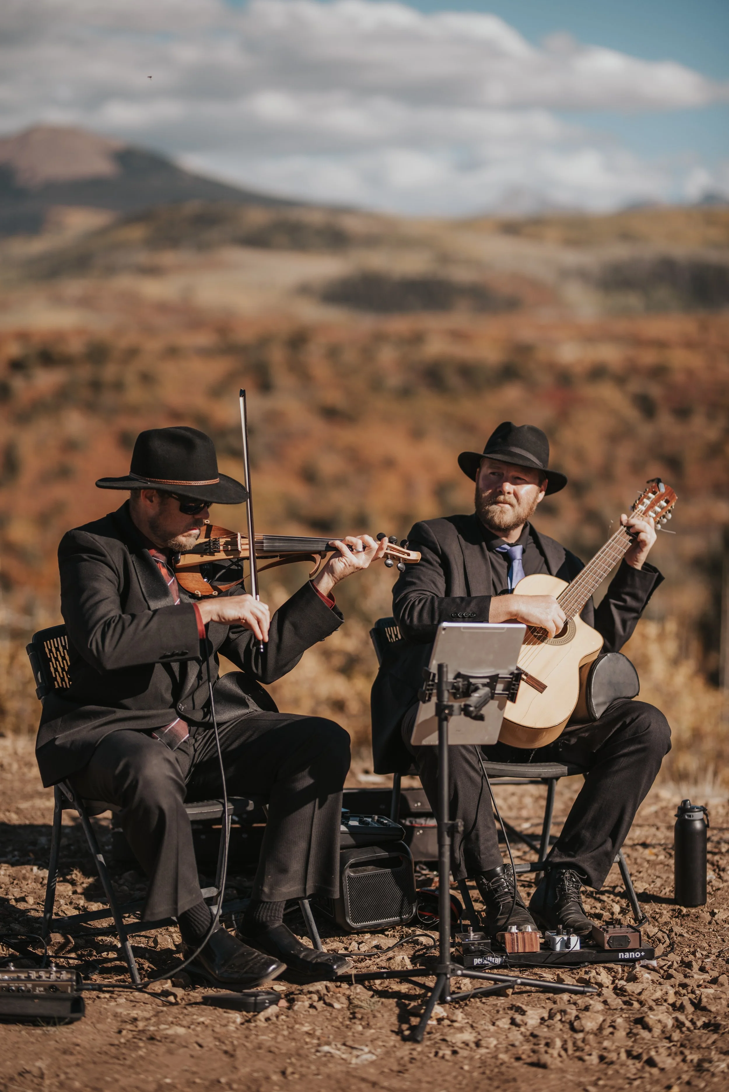 Two musicians in black suits and black hats playing guitar and violin outdoors on a rocky terrain with mountains and cloudy sky in the background.