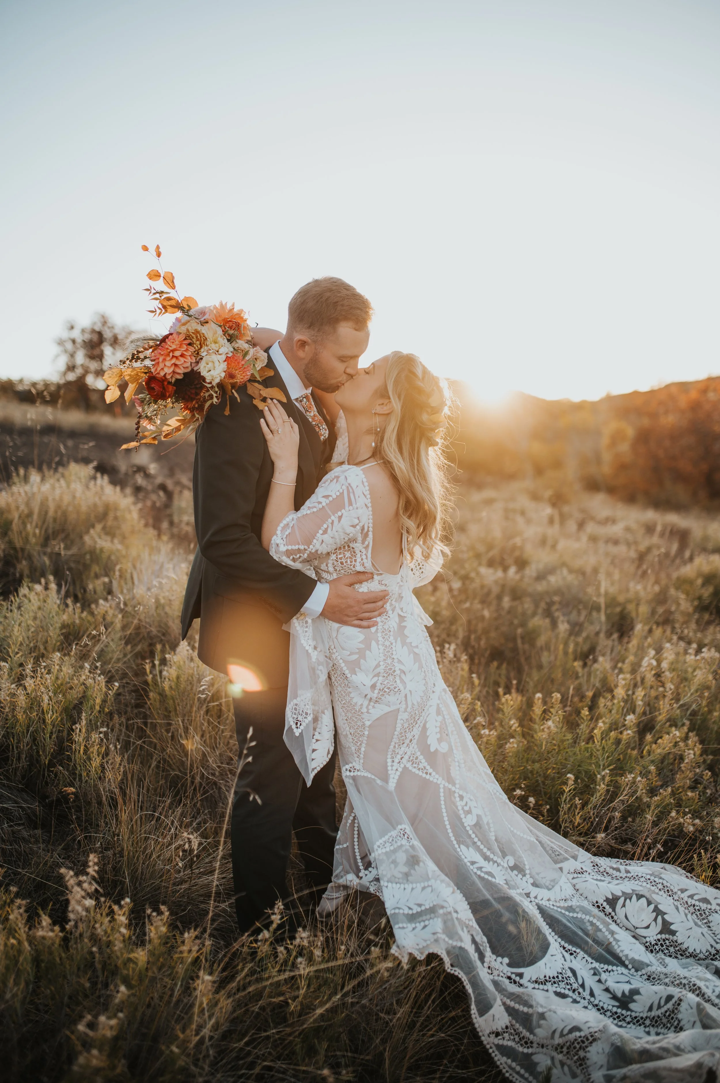 A bride and groom sharing a kiss in a field during sunset, with the bride holding a bouquet and wearing a lace wedding gown, the groom in a black suit.