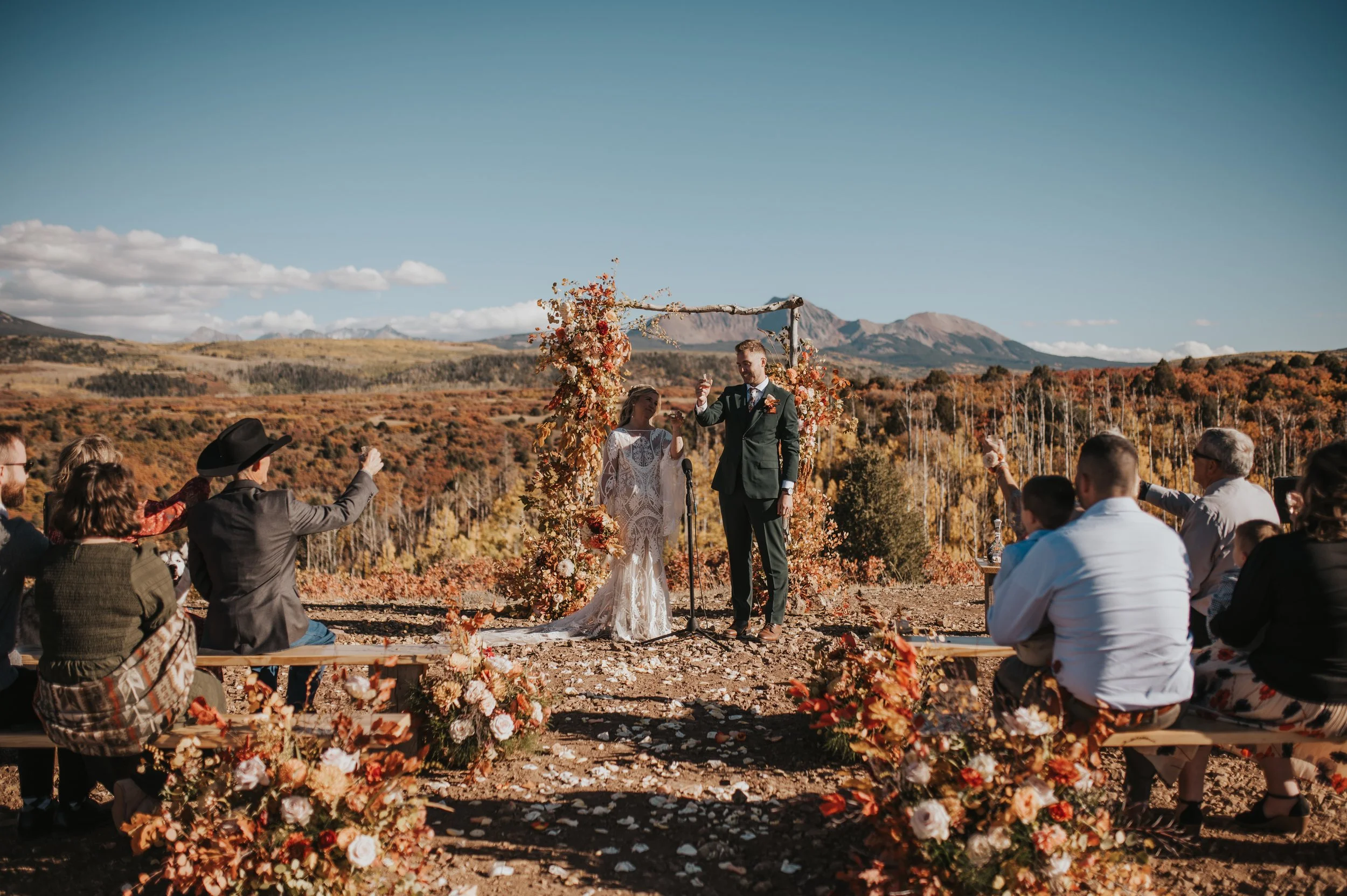 A wedding ceremony outdoors with a bride and groom standing under a floral arch, surrounded by guests, set against a scenic mountain landscape with autumn foliage.
