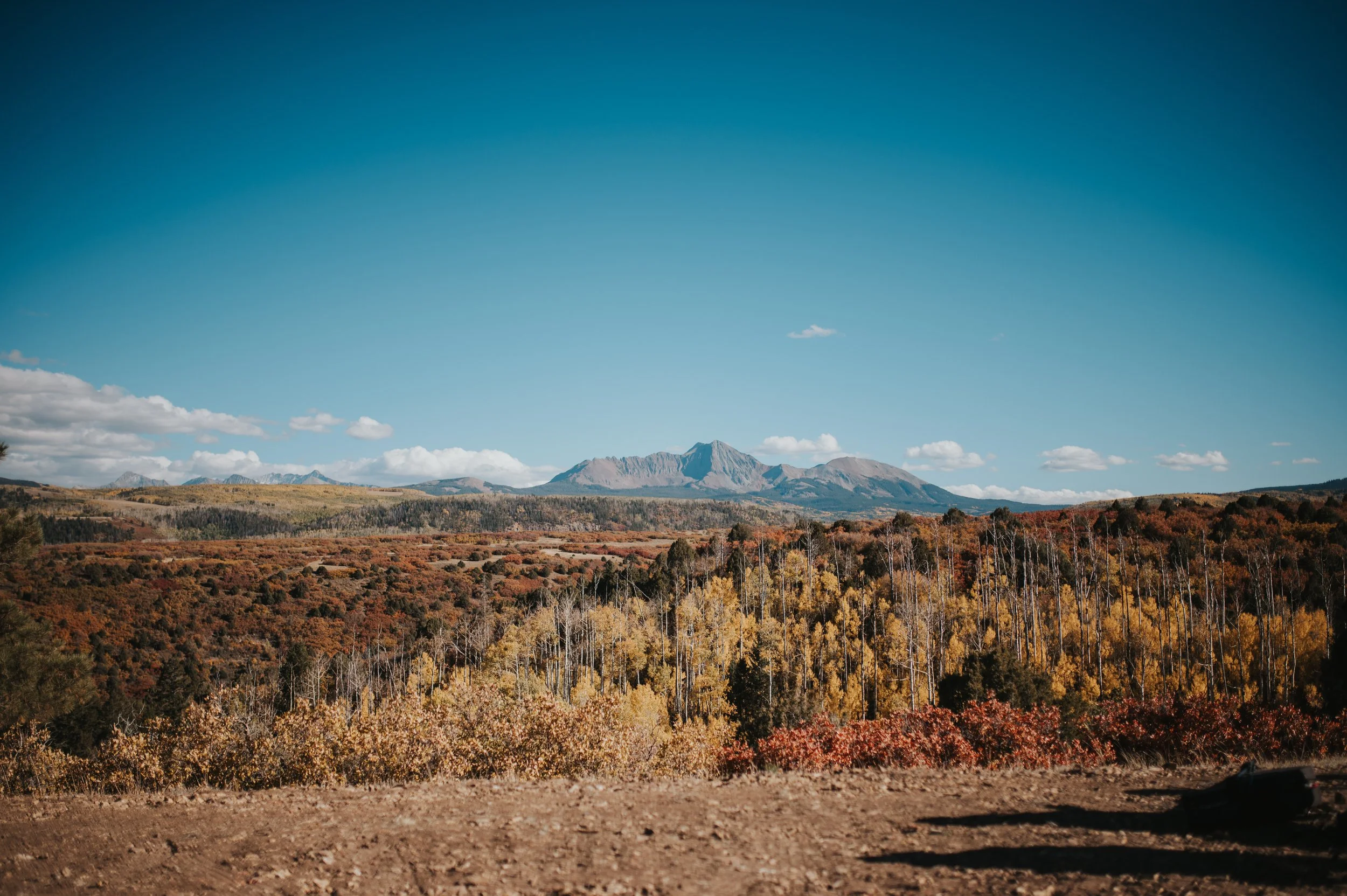 Scenic view of a mountain range with colorful autumn trees in the foreground, under a clear blue sky with scattered clouds.