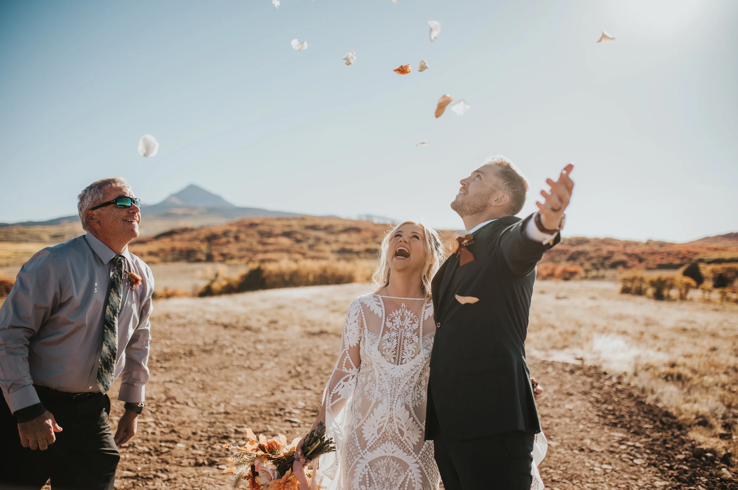 A wedding celebration outdoors in a desert landscape with mountains in the background. The bride and groom are throwing flower petals in the air, smiling and joyful. An older man stands nearby, also happy, wearing sunglasses and a tie.