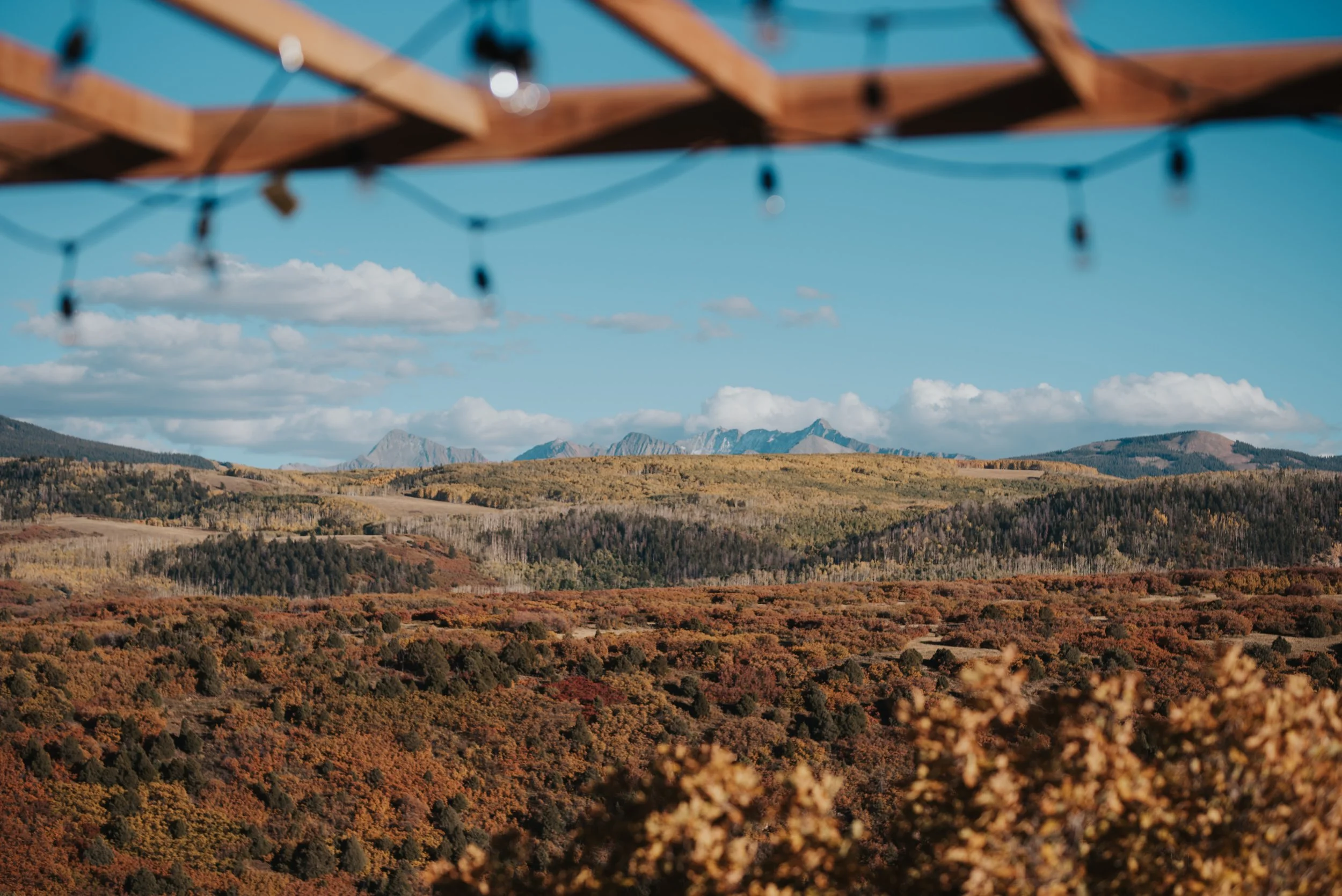 Scenic landscape of rolling hills and mountains under a blue sky with scattered white clouds, viewed through a partially blurred wooden structure with string lights.