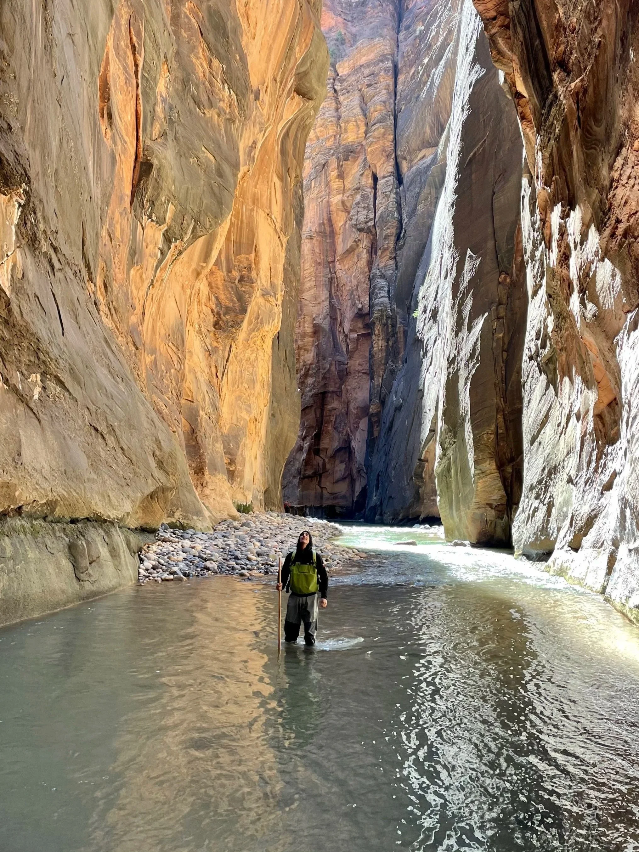 A person standing in a creek within a narrow canyon with high, colorful rock walls and a waterfall in the distance.