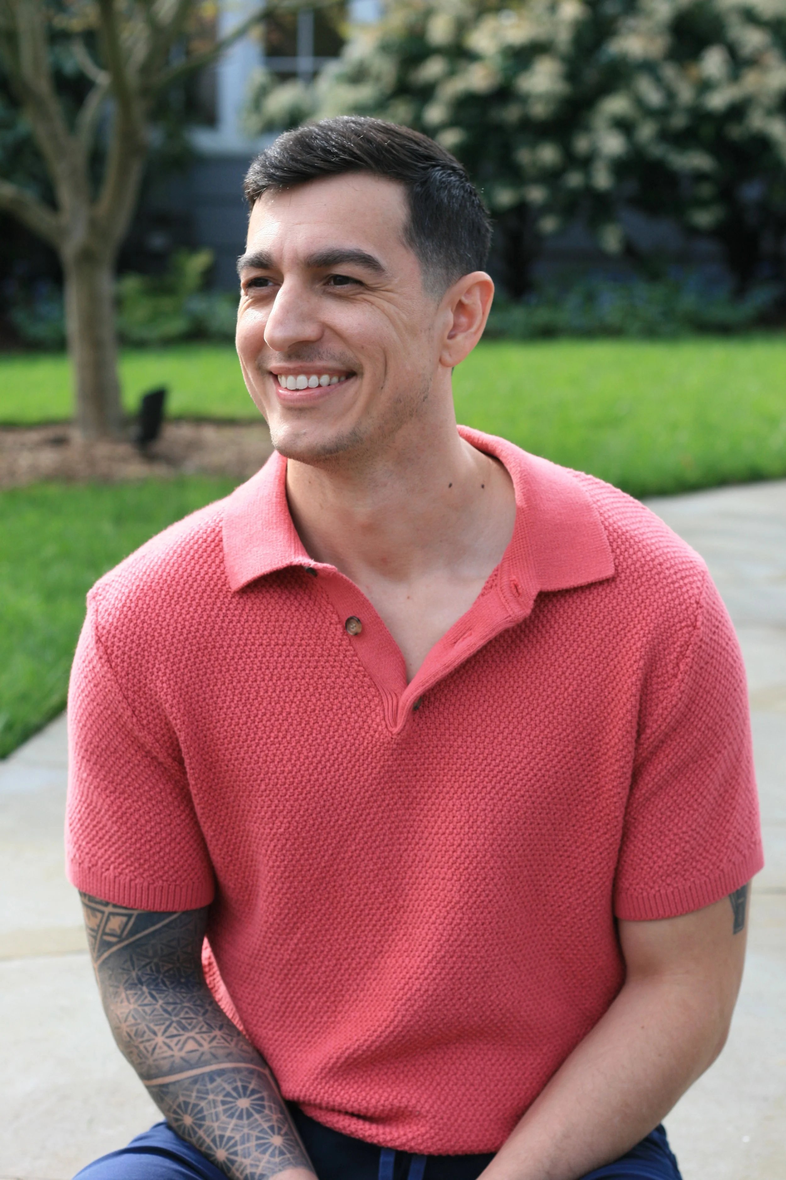 A young man with short dark hair, smiling, wearing a red textured polo shirt, sitting outdoors on a sidewalk with grass and trees in the background.