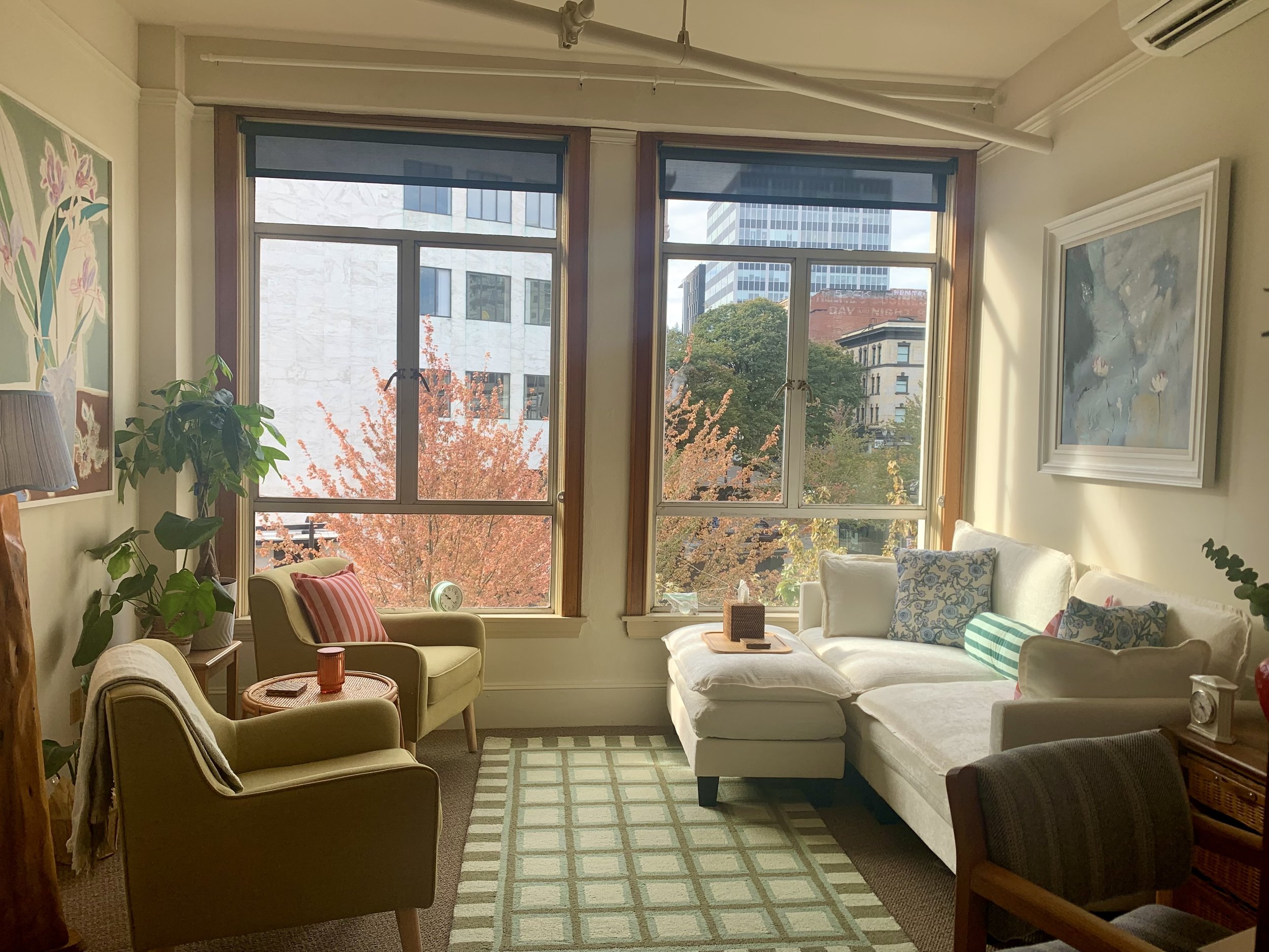 A warm office room with large windows showing city buildings and trees outside. The room has cream-colored walls, a beige armchair with a striped pillow, a white sofa with patterned pillows, a green rug, and various potted plants.