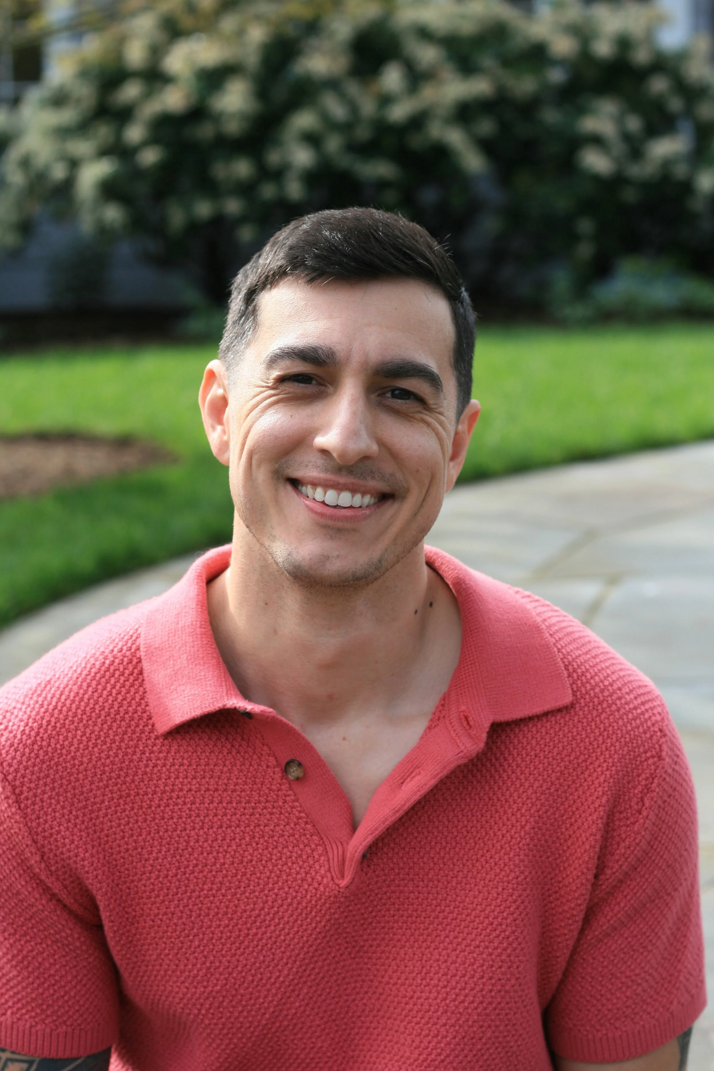 A young man with short dark hair smiling outdoors, wearing a pink polo shirt, with a grassy area and trees in the background.