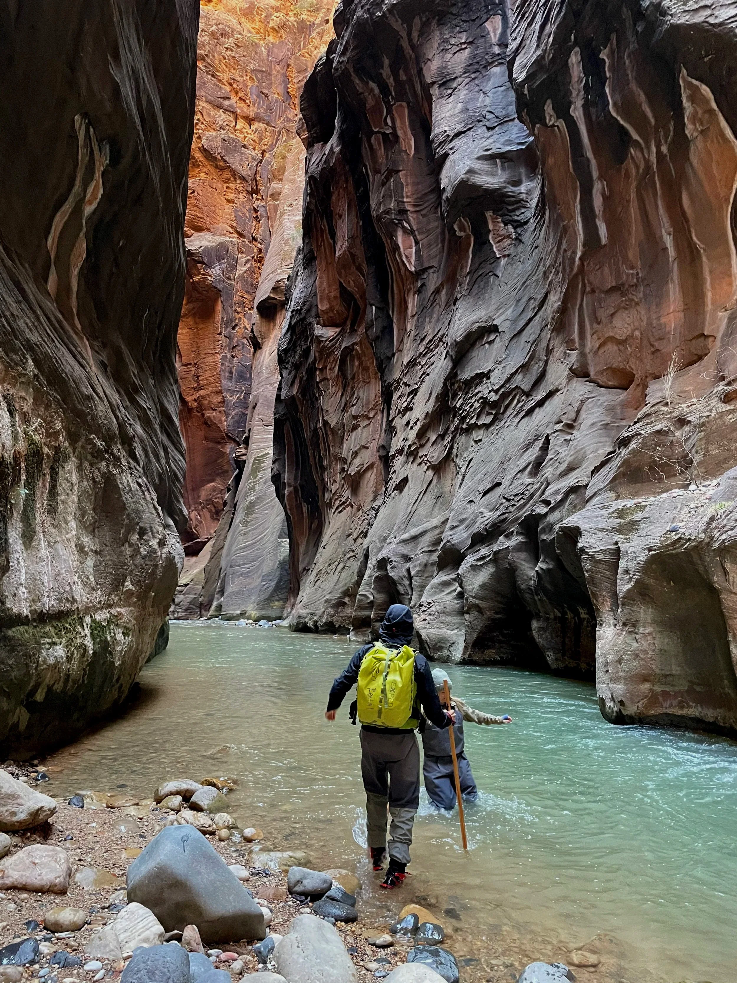 One person helping another wade through a narrow canyon with steep, reddish-brown rock walls deepening into a greenish river.