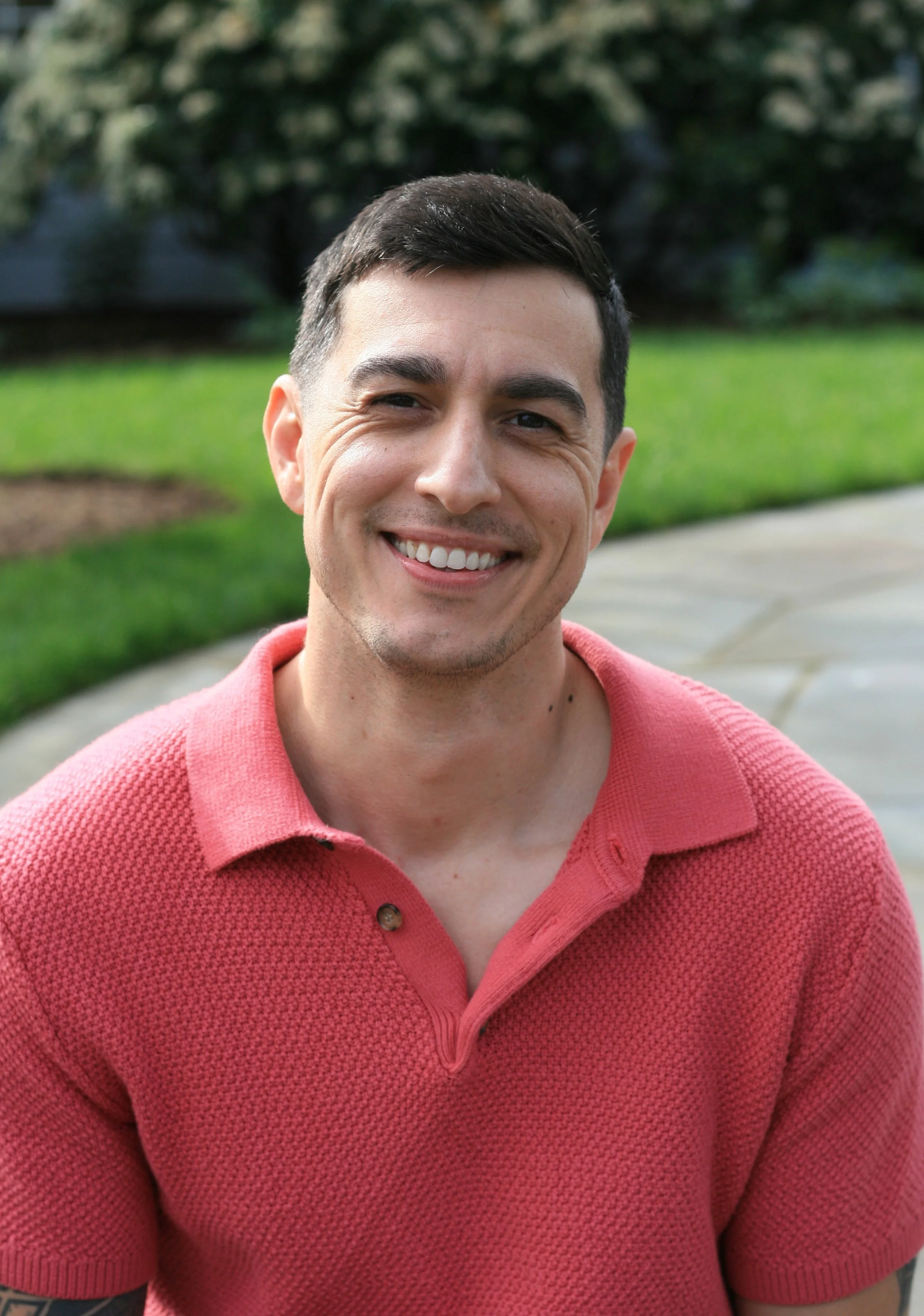 A young man with short dark hair, smiling outdoors in a park with green grass and trees in the background, wearing a red textured polo shirt.