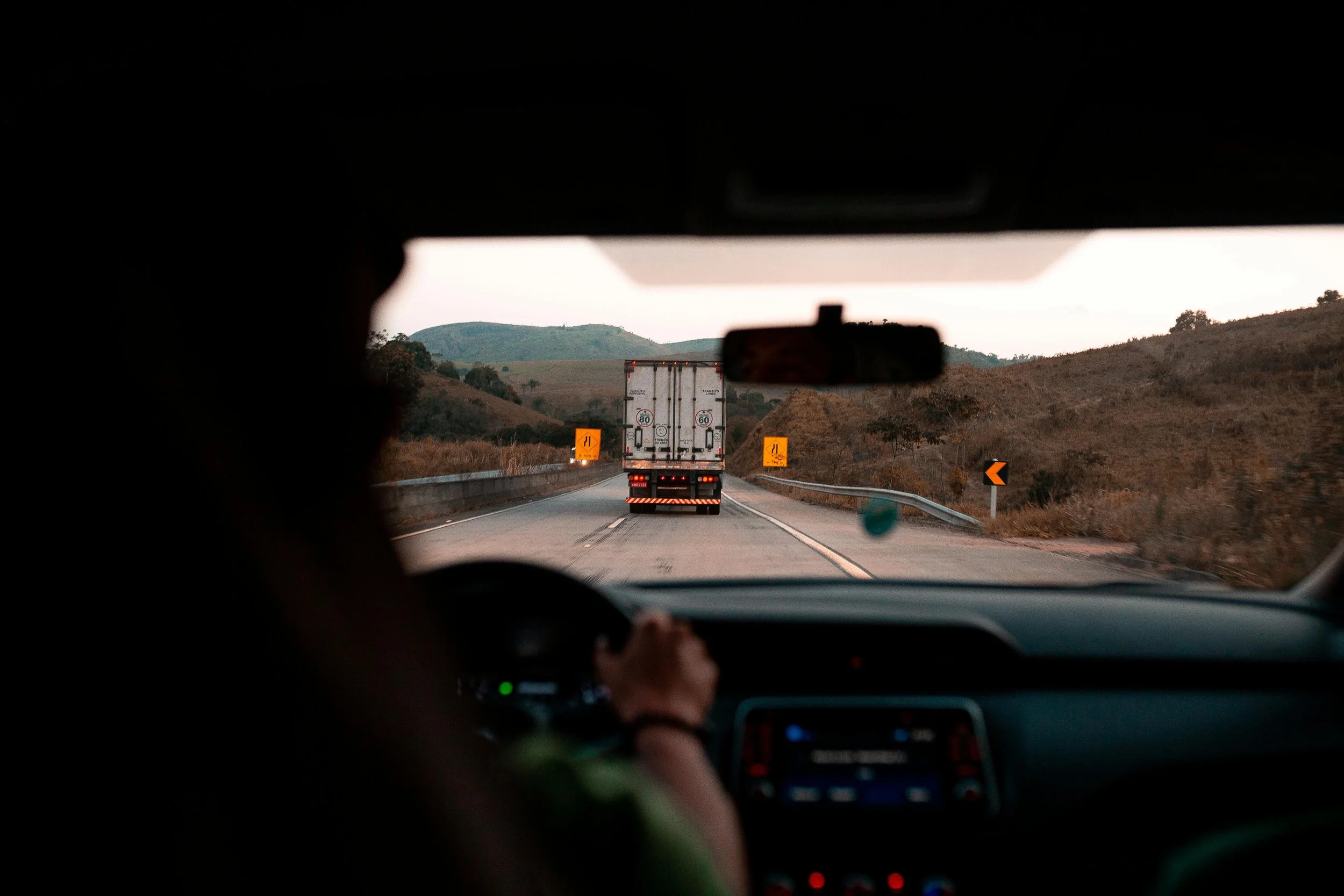 Inside a vehicle looking through the front windshield at a two-lane road with a truck ahead, surrounded by hills and landscape, traffic signs on the side of the road, and a driver with hand on the steering wheel.
