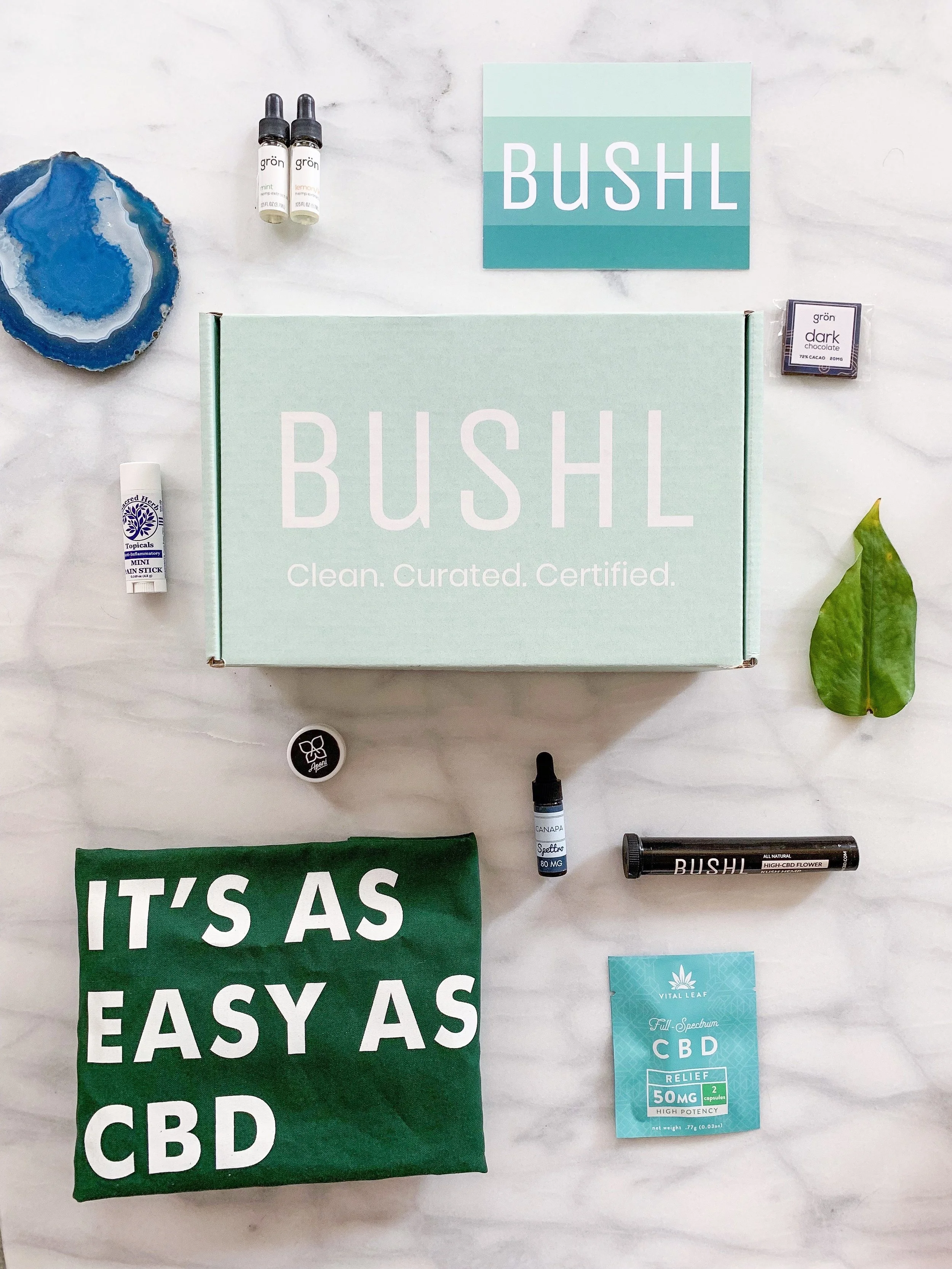 A CBD product box with various CBD-related items surrounding it, including two small bottles, a CBD spray, a green pouch with white text, a small jar, a green leaf, a bar of dark chocolate, and a CBD relief packet, all on a white marble surface.