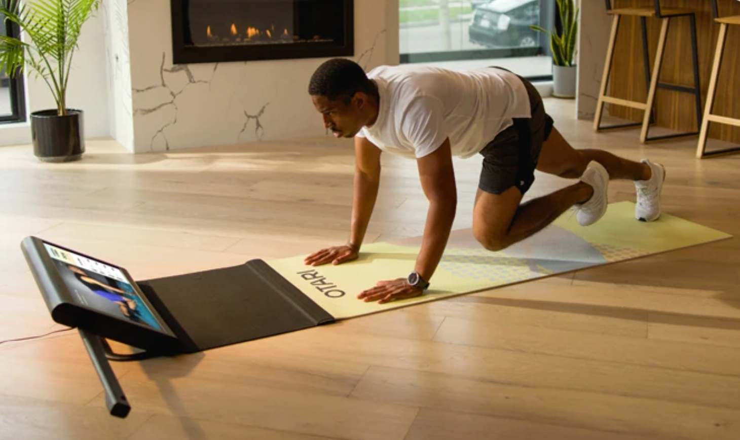 A man doing a workout on a yoga mat in front of a smartphone on a stand in a living room with hardwood floors, a fireplace, and large windows.
