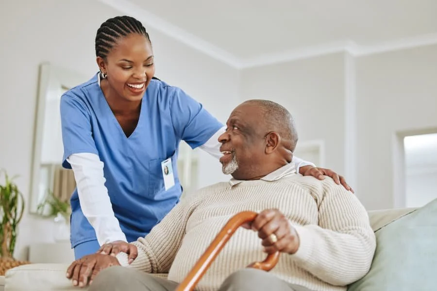A nurse smiling and talking with an elderly man sitting on a couch in a bright room, holding a cane.