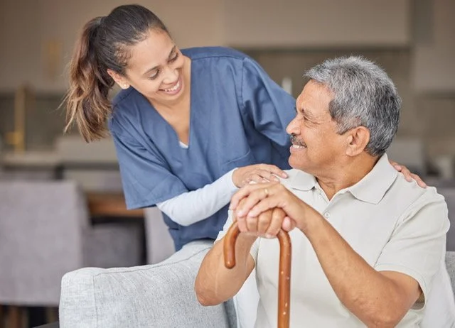 A young female caregiver talking to an elderly man with a cane, smiling in a caregiving setting.