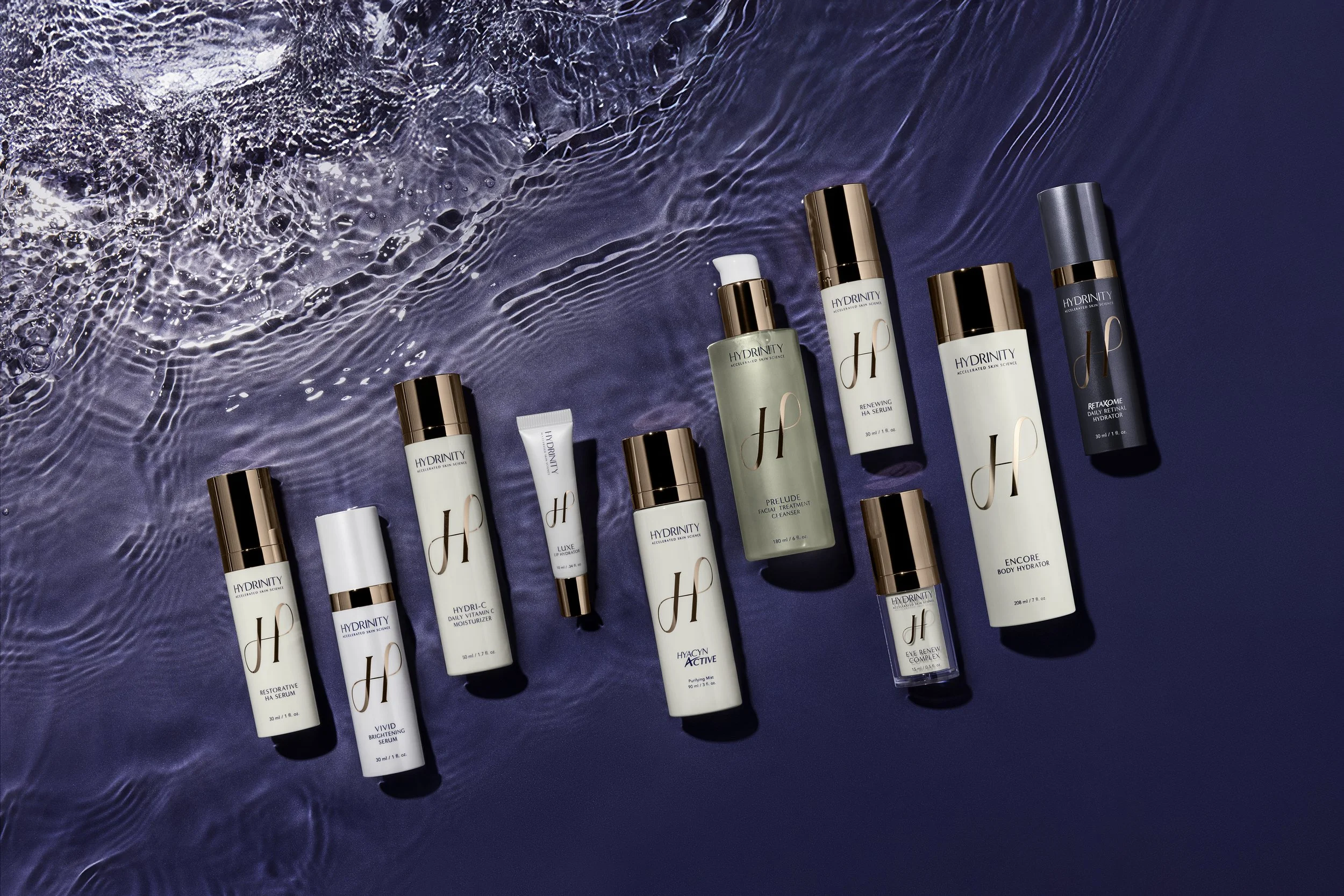 Collection of skincare products from the brand Hydrinity, arranged on a water surface with ripples, consisting of serums, moisturizers, and treatments in bottles and tubes with gold and silver caps.