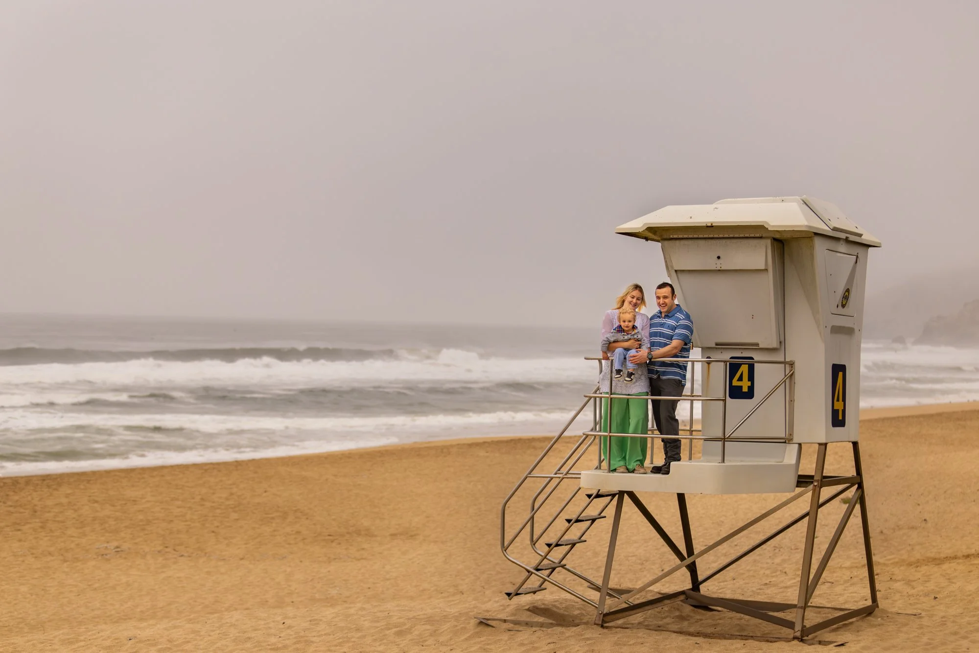 A family of three stands on a lifeguard tower at the beach, smiling and happy. The ocean waves are in the background on an overcast day.