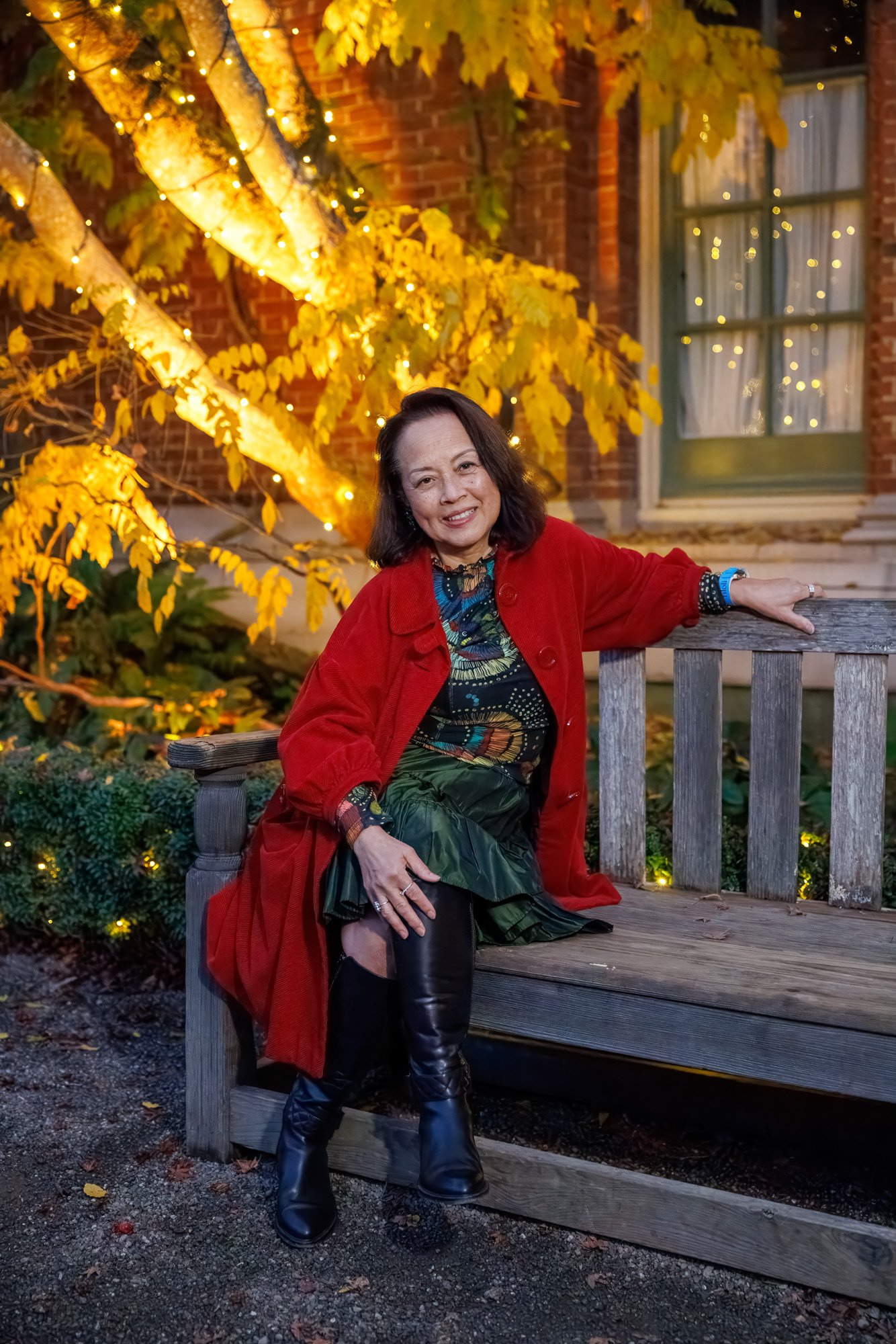 Woman in red coat sitting on garden bench beside illuminated autumn tree at Filoli Historic House, holiday portrait session Bay Area