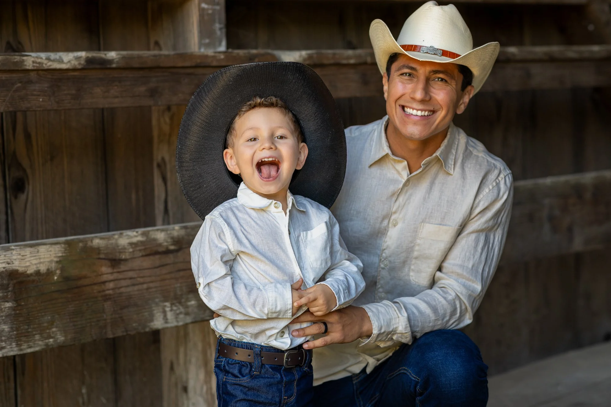 A man and a young boy wearing cowboy hats, smiling and posing outside against a wooden fence. The man is wearing a white cowboy hat and a light beige shirt, while the boy is wearing a large black cowboy hat and a light-colored shirt.