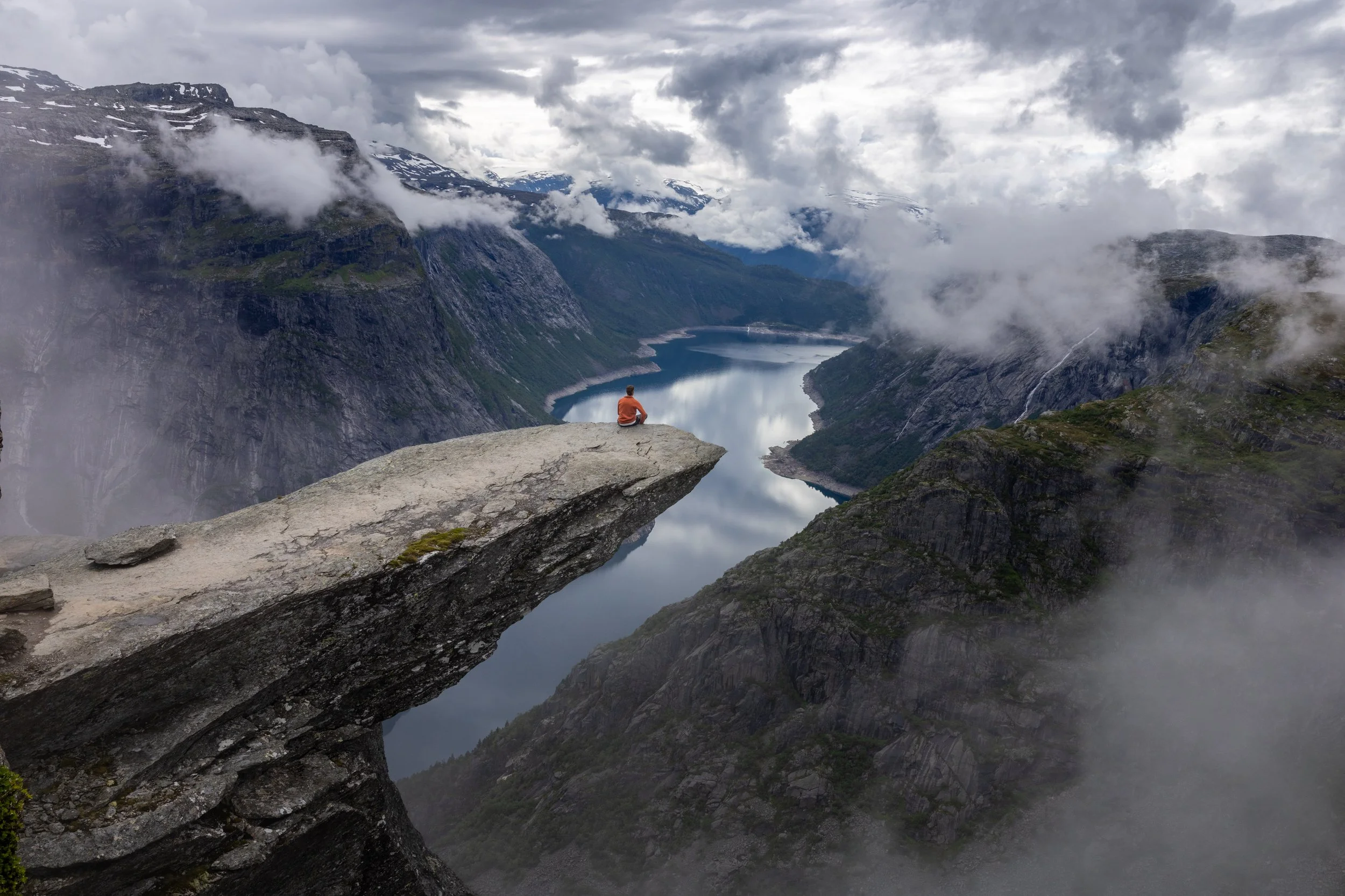 A person sitting on a large rock ledge overlooking a lake and steep mountains with clouds and fog surrounding the area.
