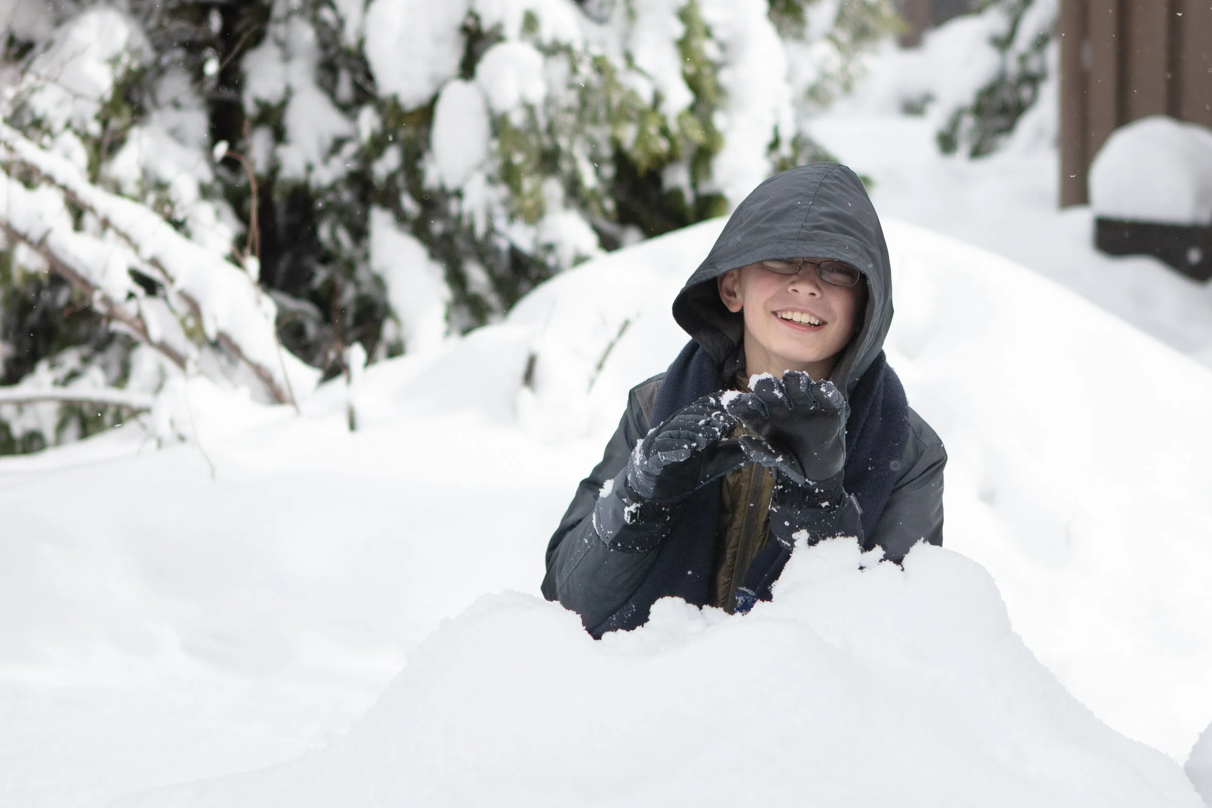 Yosemite family adventure photography session - A boy wearing a dark hoodie, glasses, and gloves playing in the snow, smiling.