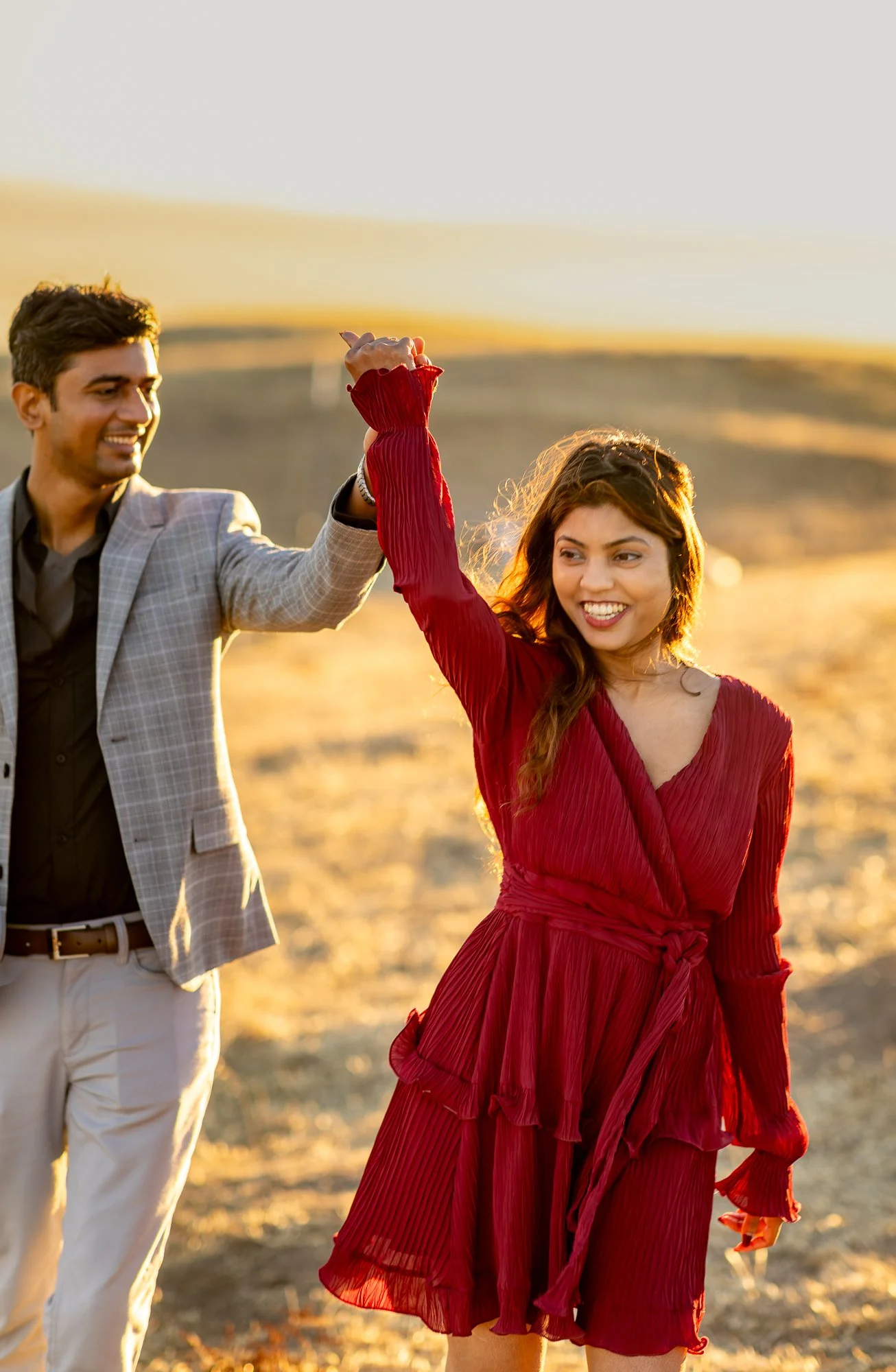 A man and woman holding hands in a landscape during sunset, smiling, with the woman in a red dress and the man in a suit with a checkered blazer.
